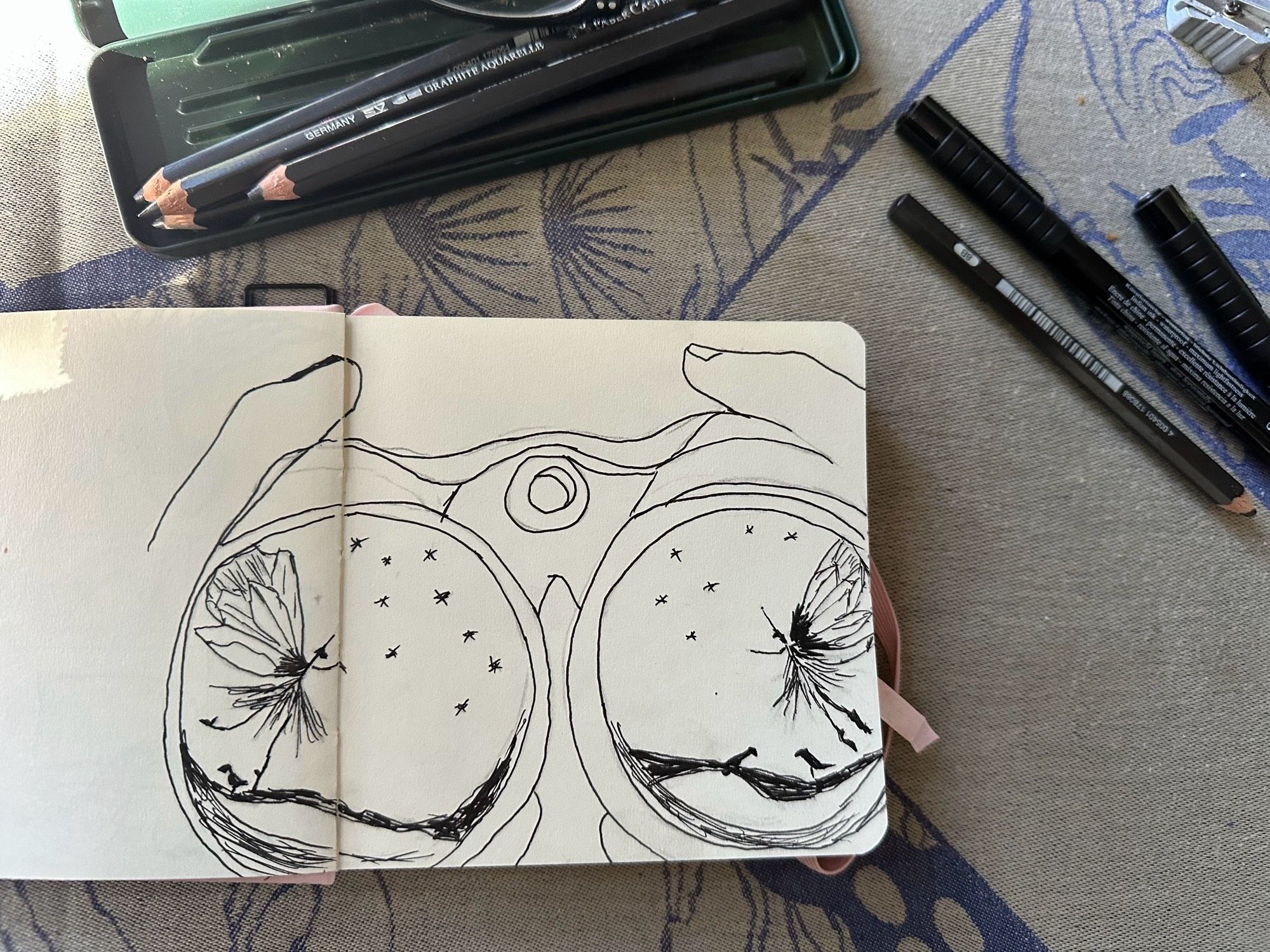 La photo montre un carnet de croquis avec un dessin de deux objets circulaires qui ressemblent à des jumelles mais une vue qui ne peut pas exister dans la réalité et qui permet de voir ce que la personne regarde, on voit dans chaque cercle une petite créature qui danse sous les étoiles sur une branche avec des silhouettes d’oiseaux à leurs pieds, elles sont très fine telles des brindilles avec de fines ailes et j’ai essayé de faire des créatures identiques pour rappeler l’autre signification du mot jumelles en français. À côté du carnet il y a des stylos et des crayons, le tout est sur une nappe grège à motifs bleus