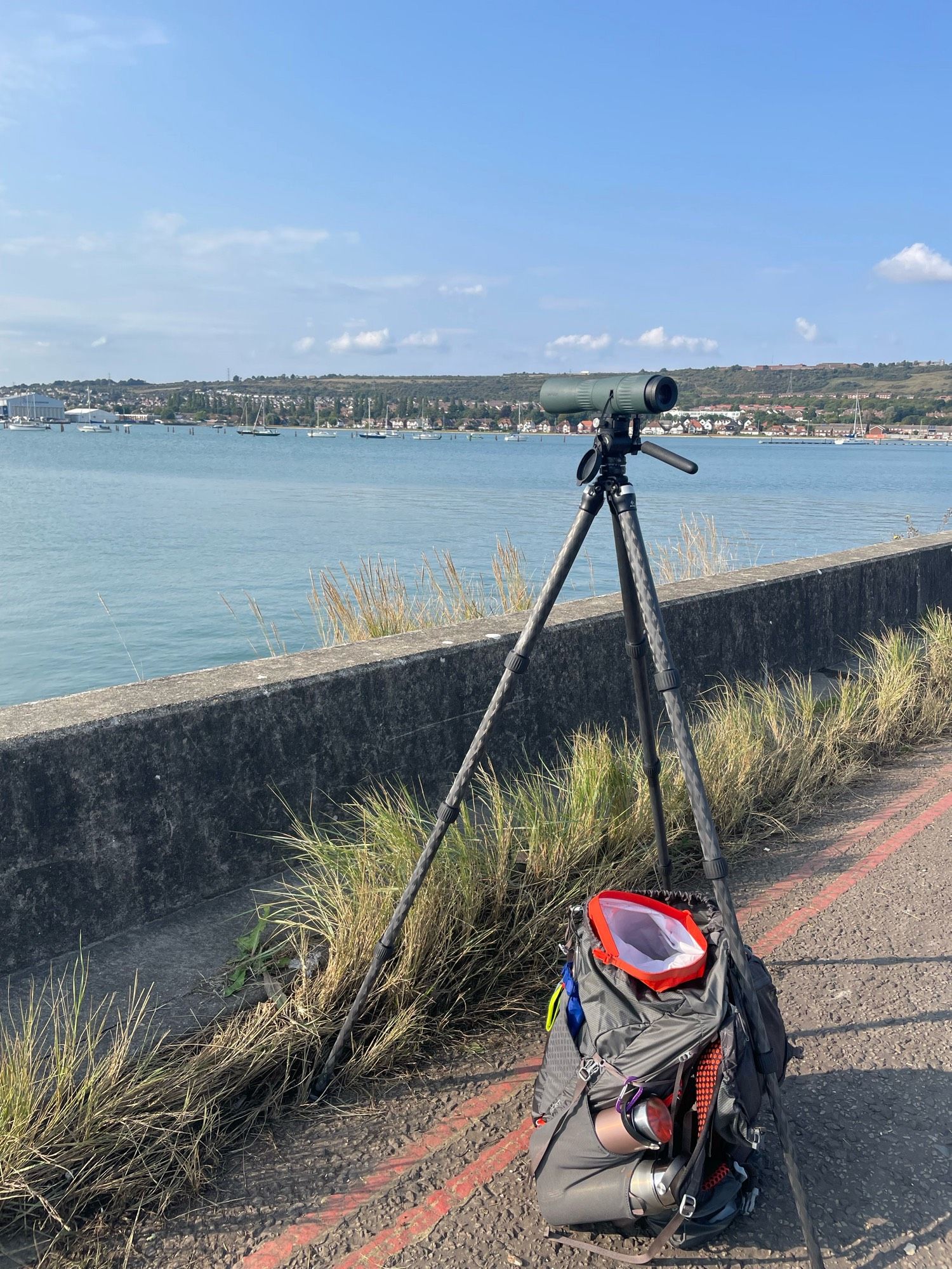 Paulsgrove Lake in Portsmouth Harbour looking towards Ports down Hill, with houses along the northern section in front of the hill. There is a tripod and telescope by the concrete seawall and a grey rucksack. It is sunny and calm