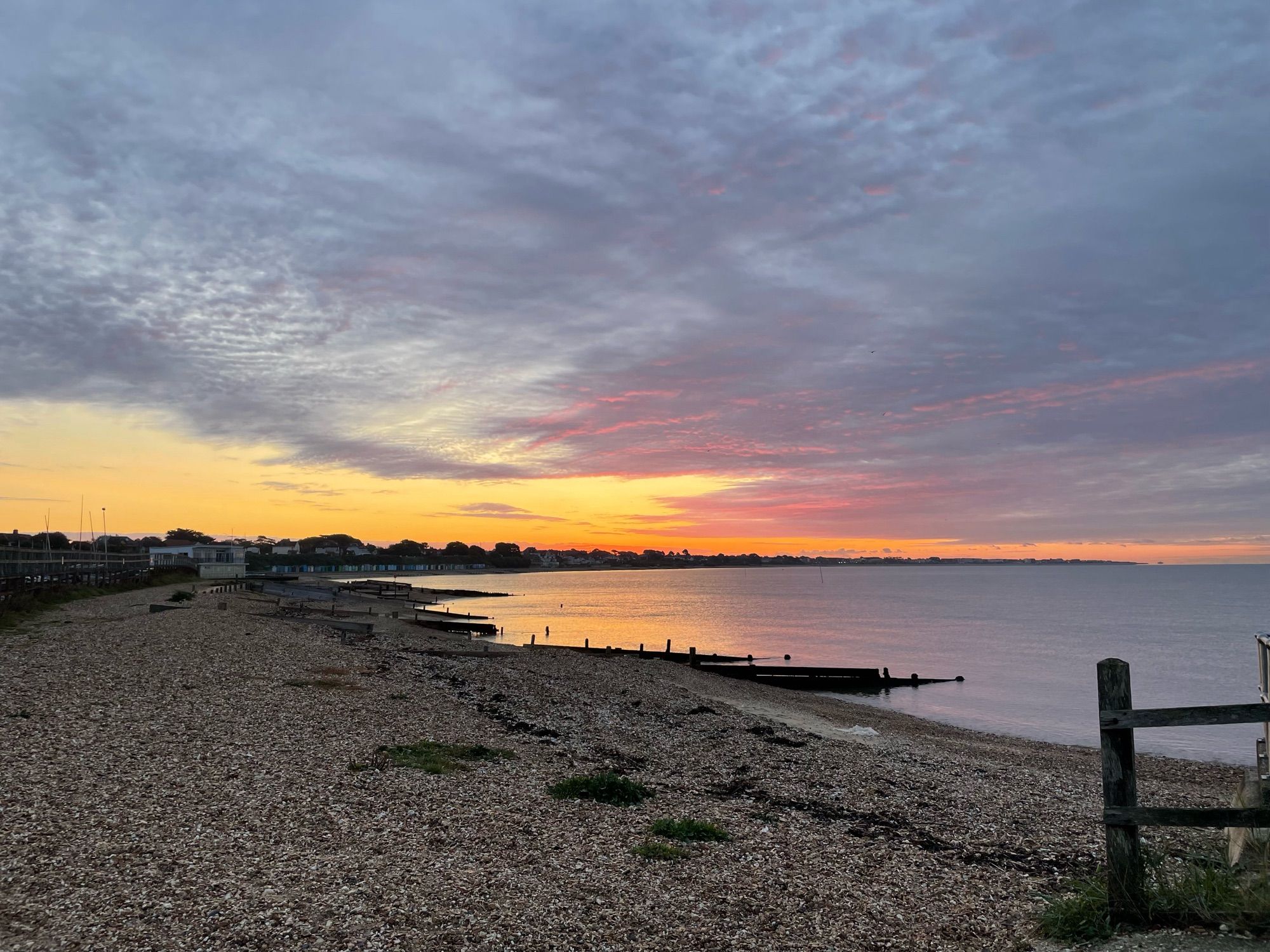 Sunrise over Hill Head. In the foreground is a shingle spit behind the harbour. The poles for sails can be seen. Behind and along the shore are beach huts below a cliff and houses above. It is mostly cloudy with some reddish clouds nearest the east. The sky is orangey yellow