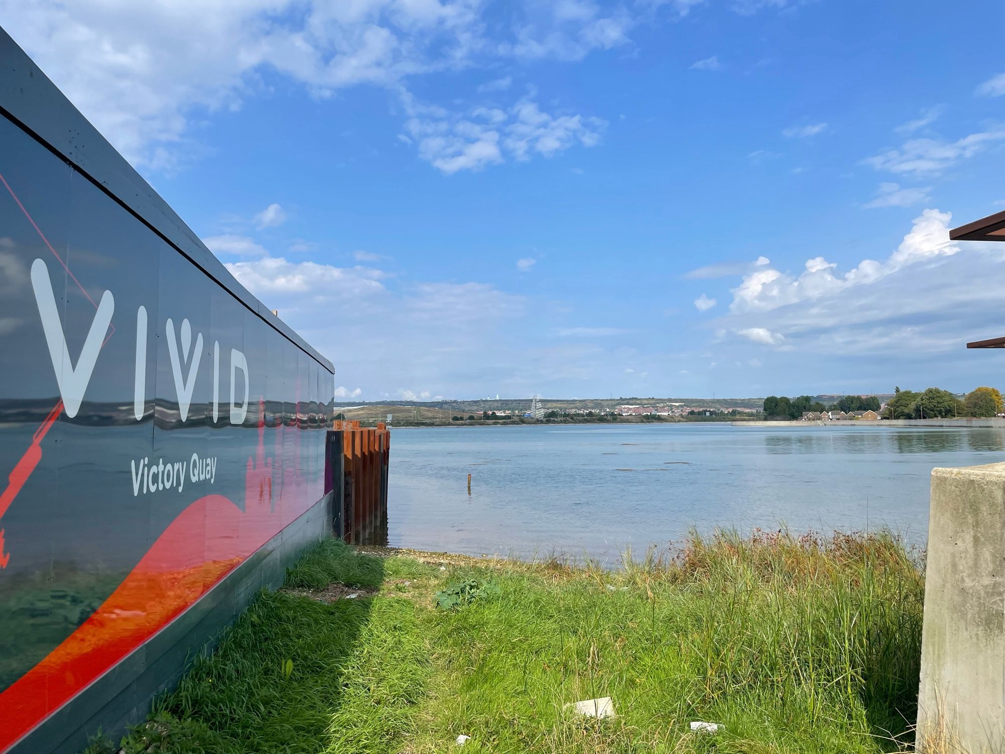 Tipner Lake by the ongoing housing development called Victory Quay. There is some grass infront and part of the concrete seawall is visible. It is sunny and calm