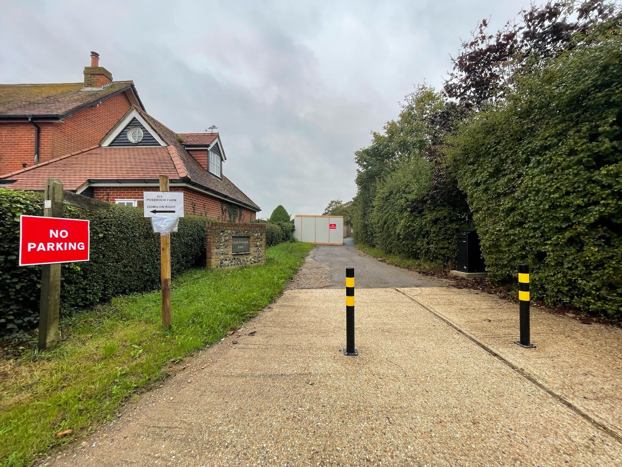 Entrance to Great Posbrook Farm. There are black and yellow bollards blocking access to vehicles and a red sign with "no parking". Entrance is between a house and hedge. There is grey boarding hiding a large pipe