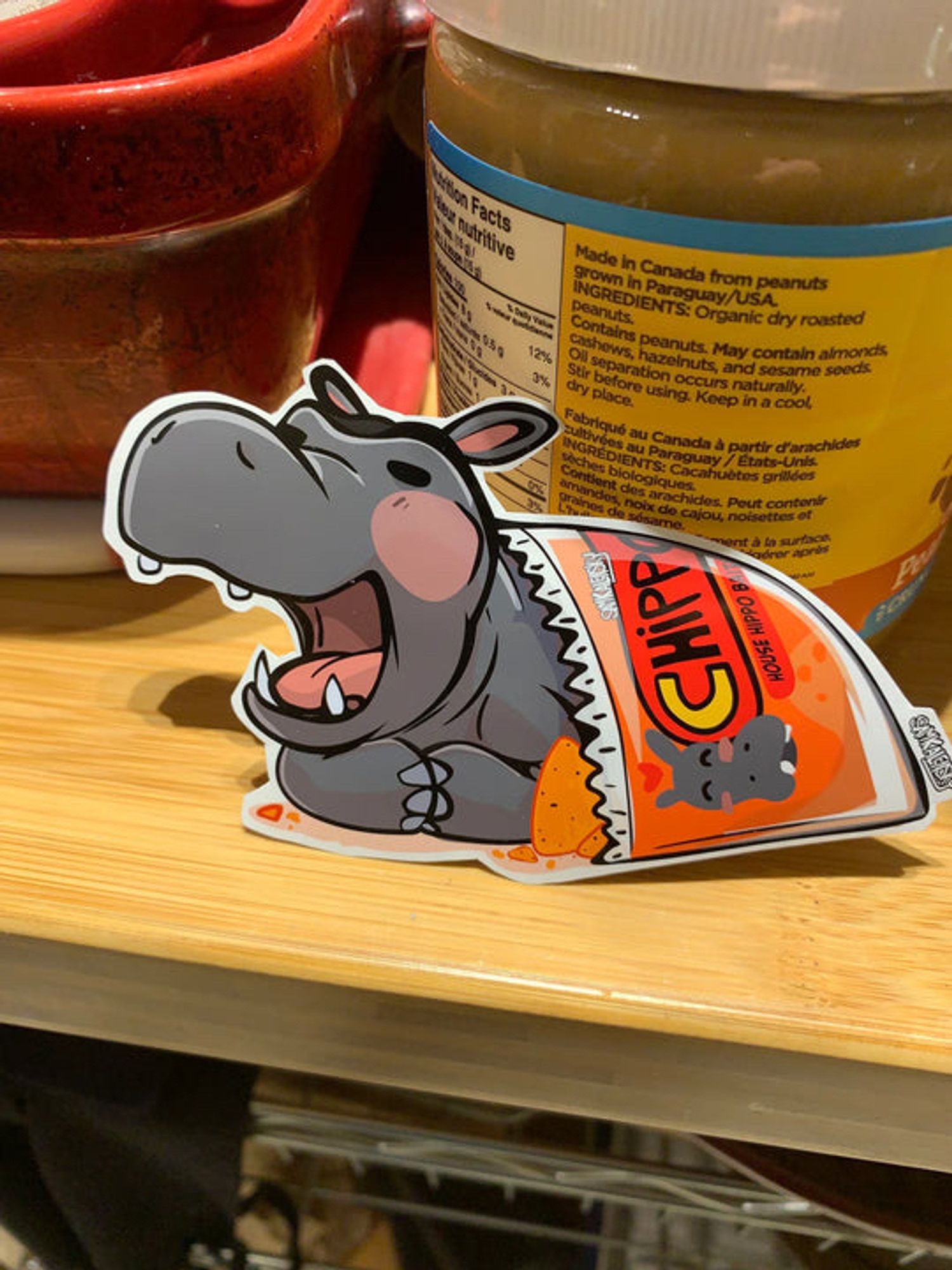 a tiny hippopotamus lets out a YAWN, its seems to have enjoyed its meal of chips and is falling asleep, theres a peanut butter jar in the background for scale. sticker art by samkalensky