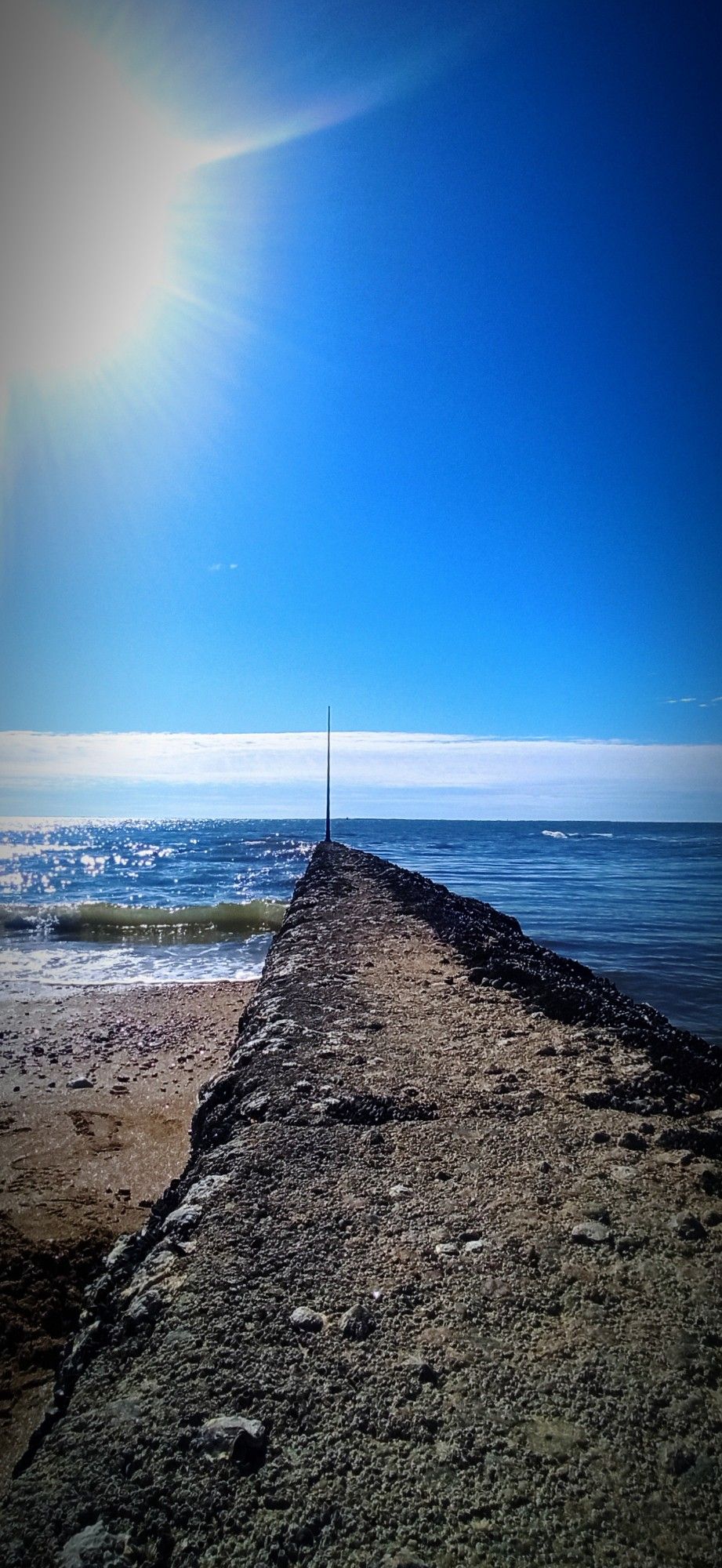 Vue sur la mer depuis une plage. Au centre de la photo une digue sépare la mer en deux parties distinctes. Le soleil brille en haut à gauche dans un ciel bleu avec une bande de nuages à l'horizon.