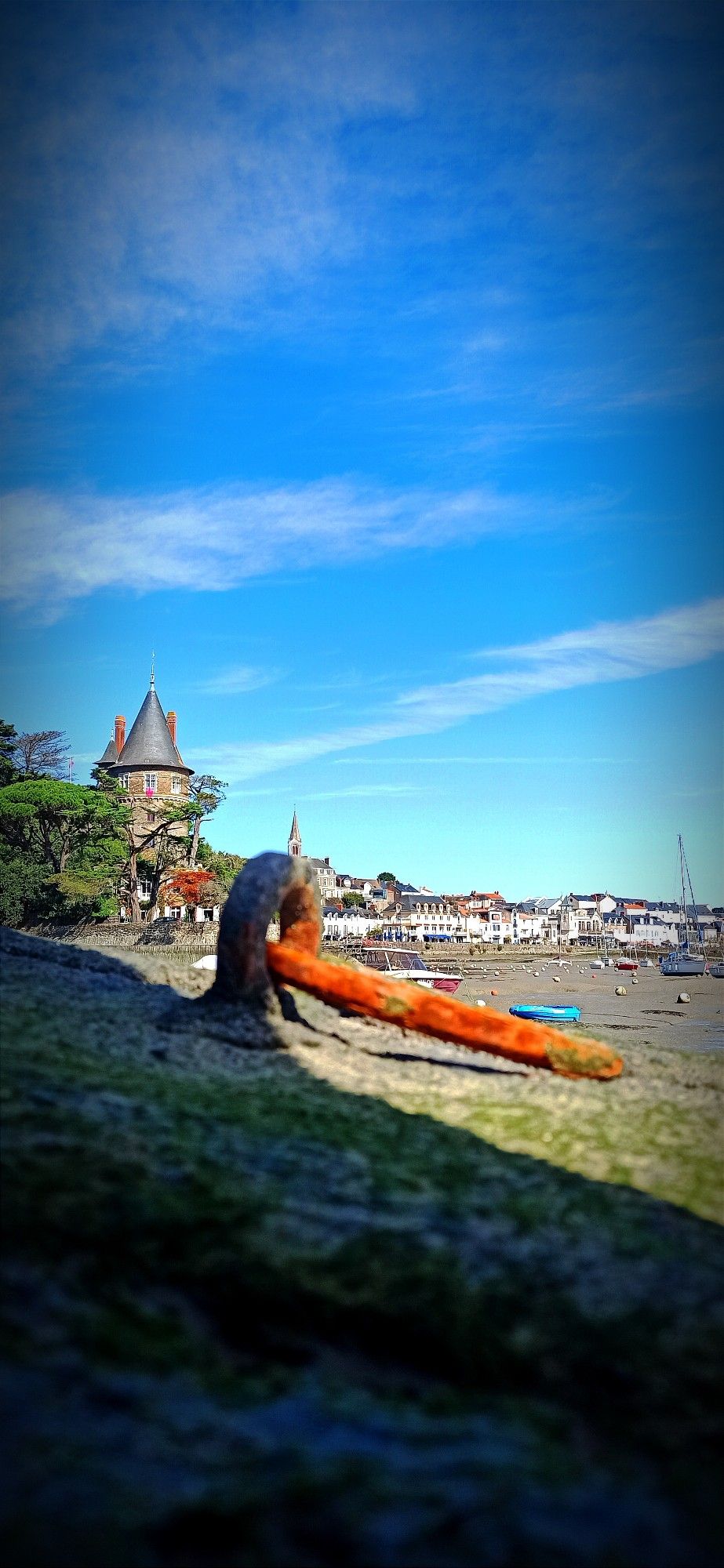 Port de Pornic à marée basse. Au loin on distingue les bâtiments et sur la gauche le château de la ville. Les bateaux sont posés sur la vase. Au premier plan, un anneau d'amarrage rouillé.