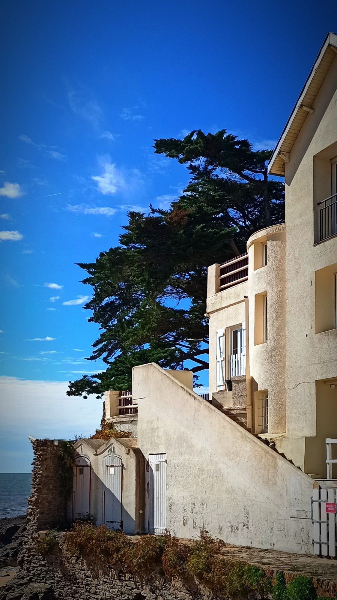 Maison de bord de mer de couleur crème. La maison est construite sur plusieurs niveaux et trois cabines de plage sont visibles au pied du bâtiment. En arrière plan derrière la maison, un arbre se dresse devant un ciel bleu.