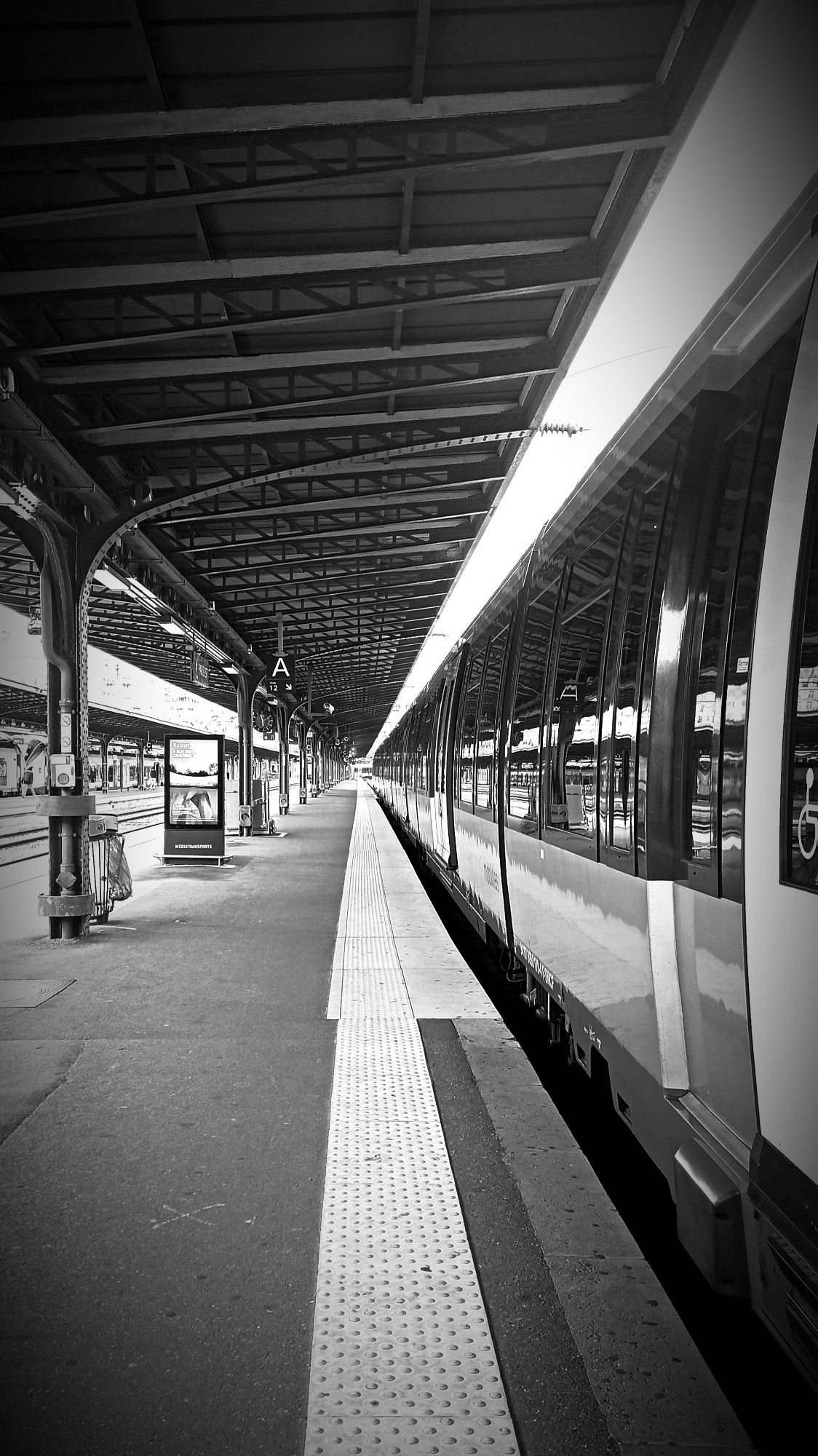 Cette photo en noir et blanc montre un quai de gare, avec un train à droite prêt à partir. La perspective plongeante, avec le toit du quai et le train qui s'étendent à perte de vue, accentue une impression de profondeur. L'architecture métallique du quai, combinée aux lignes géométriques du train et du chemin, donne une ambiance à la fois industrielle et nostalgique. La gare semble vide.