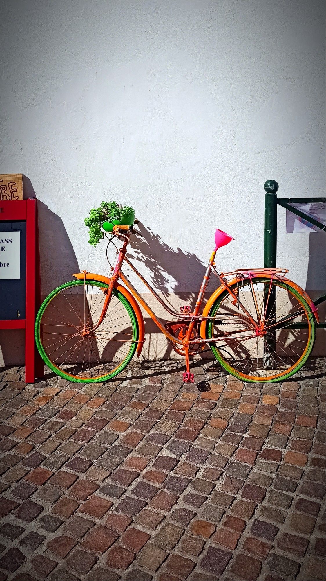 Devant la façade d’un petit théâtre nommé "Le Poche", une pancarte rouge annonce une masterclass de théâtre et chant, qui aura lieu les 4, 5 et 6 octobre. À côté de cette pancarte, un vélo coloré est adossé contre le mur blanc. Ce vélo attire l'attention par ses couleurs vives : les pneus sont peints en vert et orange, avec des touches de jaune sur les jantes. Le cadre est également dans les tons orange avec des touches de peinture, tandis qu'un pot de plante vert, installé dans le panier à l'avant, rajoute une touche de verdure. Un entonnoir rose remplace la selle. Le contraste entre les couleurs du vélo, la pancarte rouge, et le mur blanc rend la scène particulièrement vivante et originale.