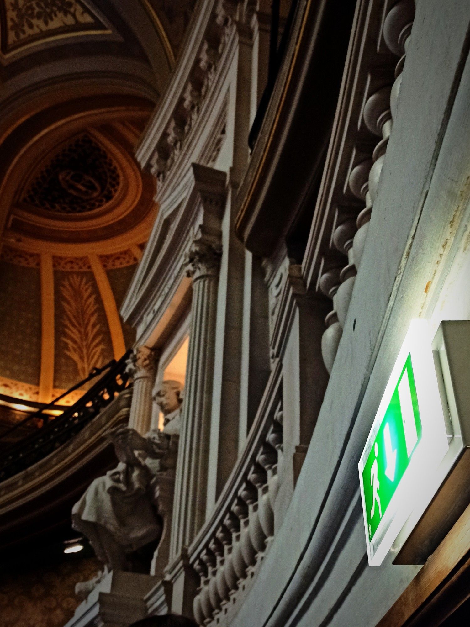 À droite, un panneau de sortie de secours lumineux vert indique une direction avec un pictogramme représentant une personne en train de sortir. Ce panneau contraste fortement avec l'architecture somptueuse et détaillée à gauche, où l'on peut voir une statue en pierre nichée dans une alcôve ornée de colonnes et de moulures complexes.

Le bâtiment présente un style néoclassique, avec des détails architecturaux riches tels que des balustrades, des frises et un plafond voûté magnifiquement décoré. La statue est celle de Pascal, le lieu étant le grand amphithéâtre de la Sorbonne.