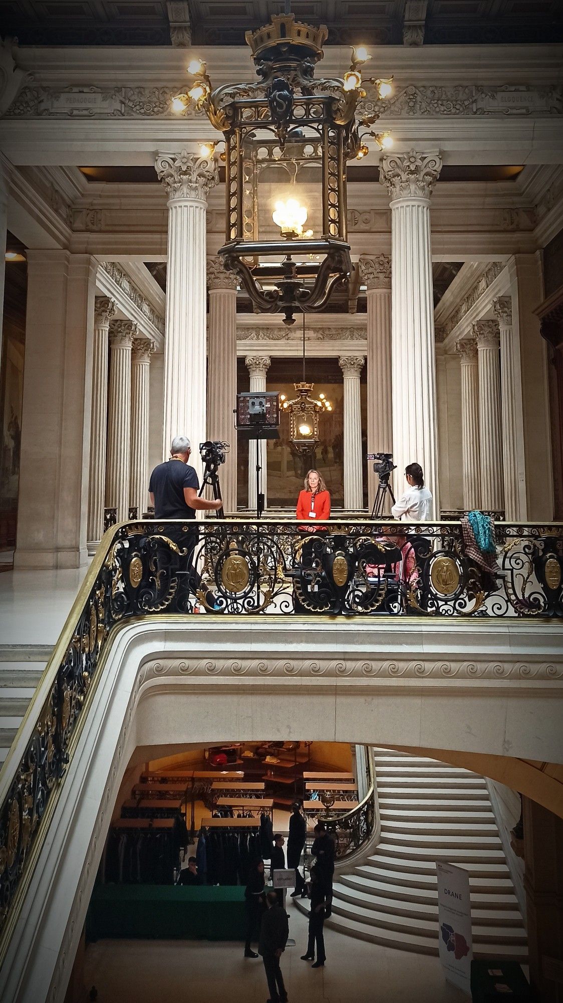 L’image montre l’intérieur de la Sorbone . Il y a un grand escalier orné avec une balustrade en fer forgé menant à un niveau supérieur où se trouvent trois personnes. Une personne en veste rouge est interviewée et filmée par deux autres personnes avec des caméras sur trepieds.  En bas de l’escalier, il y a plus de personnes et d’activités (vestiaire temporaire).