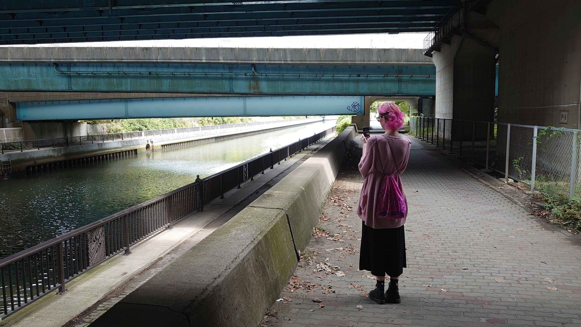 Image of a walking path under a bridge next to a canel. The image is in stark blues and greys. On the right side a person wearing boots and black pants with a pink oversized cardigan and pink hair is framed by the background