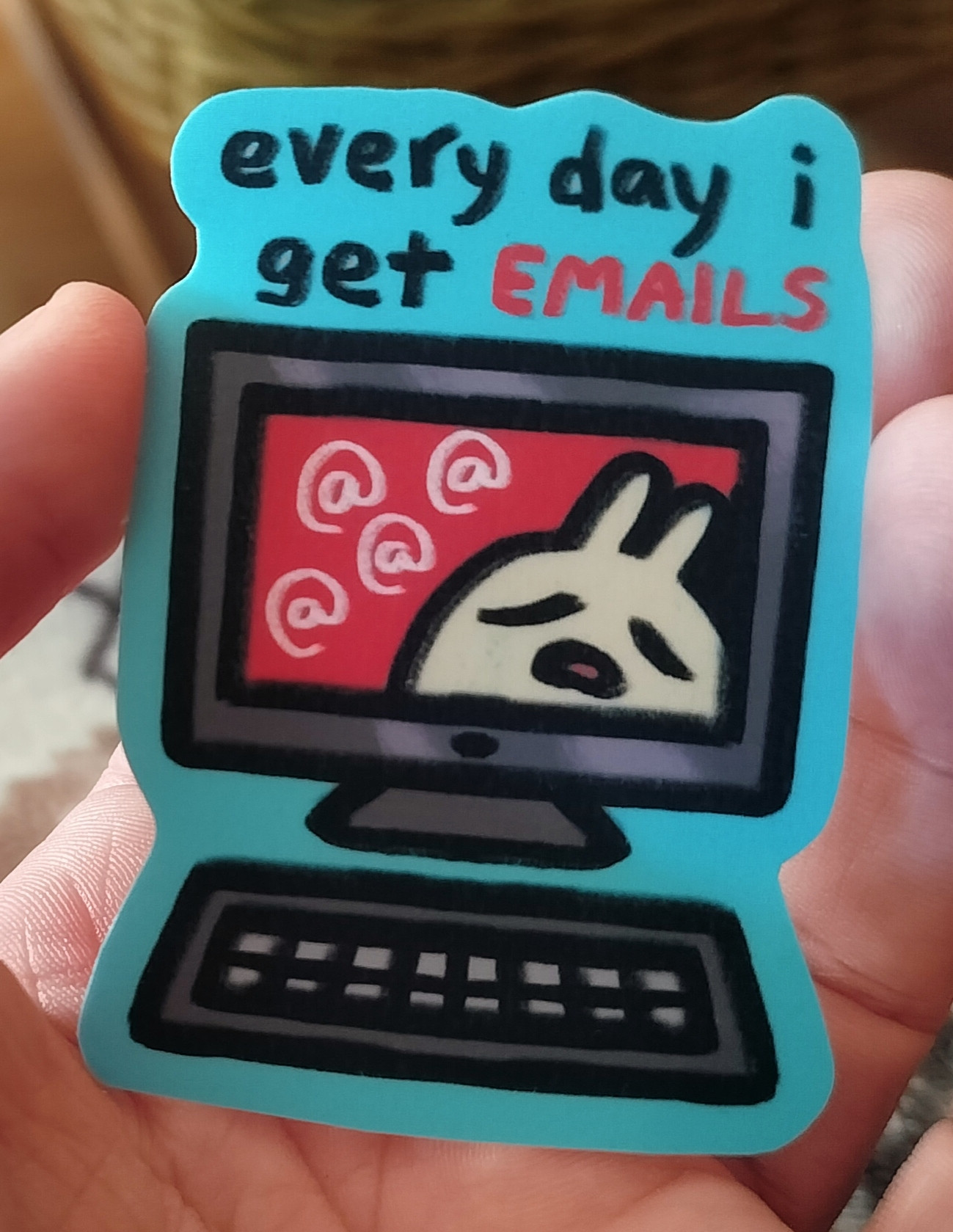 Photo of a sticker that has a dark grey PC monitor and keyboard on a bright light blue background. There is a bone white bunny on the screen with a red background and there are 4 at-signs there. The bunny looks very pensive and defeated. There is a text "every day i get emails" on top of the picture.