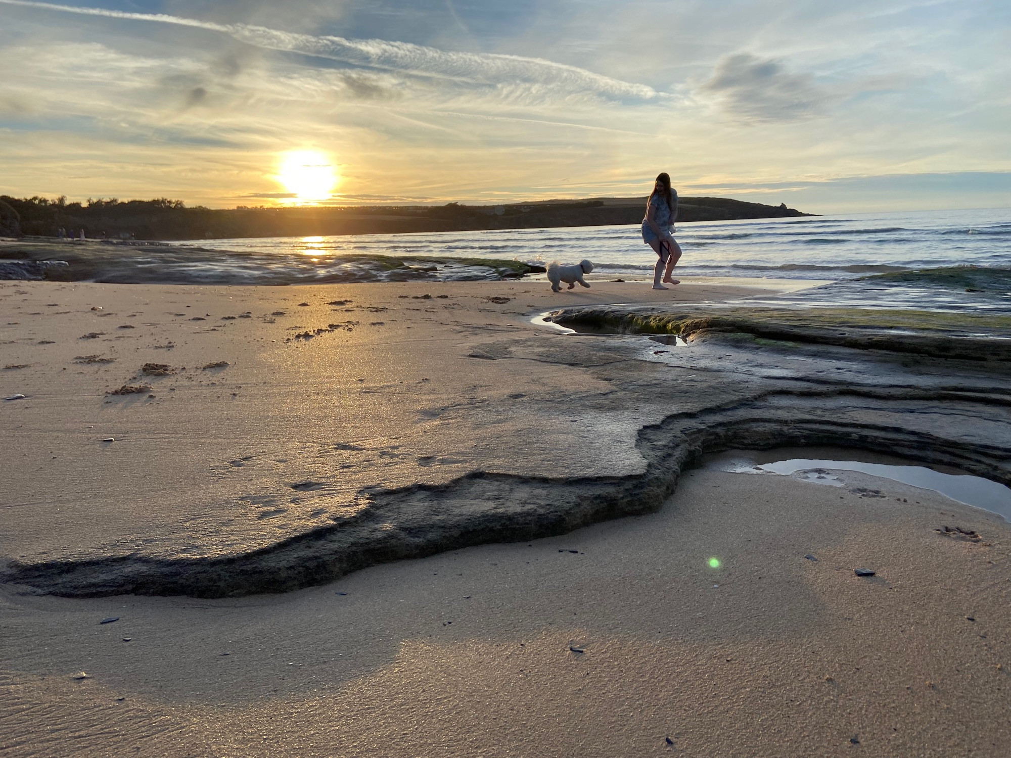 Sunset in Cornwall, Harlyn Bay. A teenager walks across the beach, sea to the right, silhouette of headland behind and small Bichon frisée at her feet