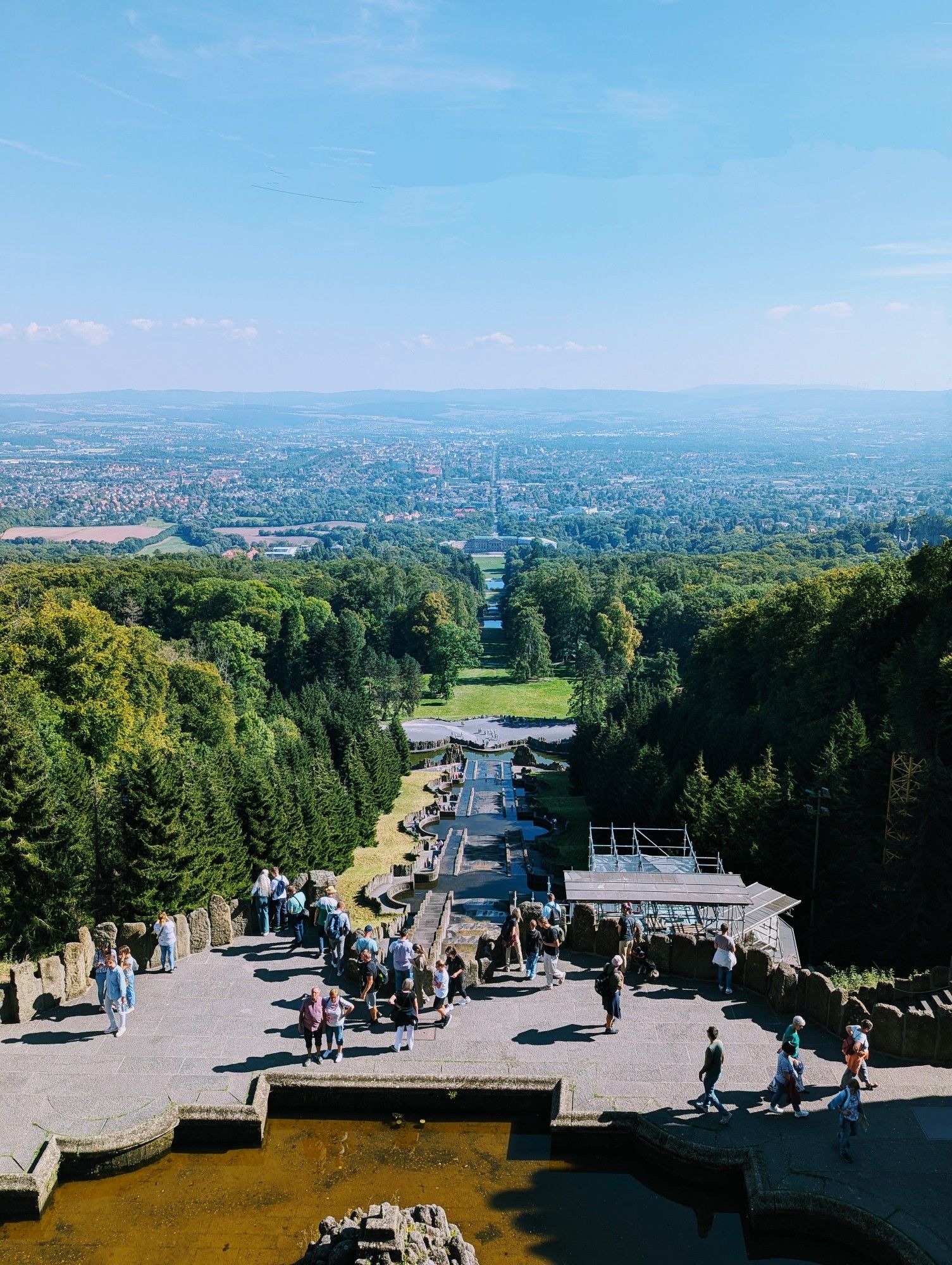 A wide angle shot of the landscape from the top of a mountain. There's a man-made cascading water feature leading straight down and away from the camera in the centre, framed by thick forest to the left and right. In the distance is a sprawling city and even further rolling hills.