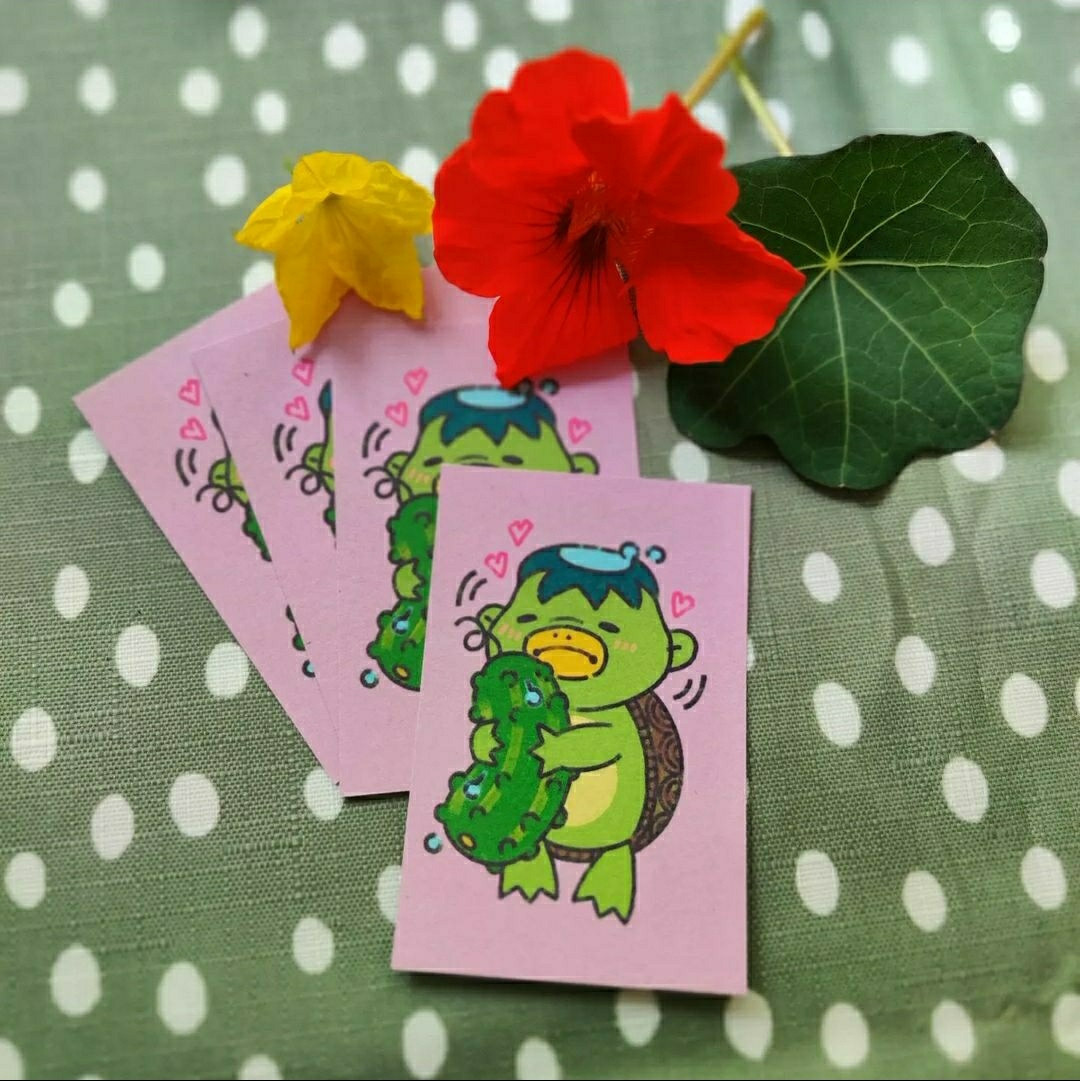 Light pink business cards with a cutified kappa beast and a cucumber on them. Red and yellow flowers as a decoration on a dotted green tablecloth in the background.