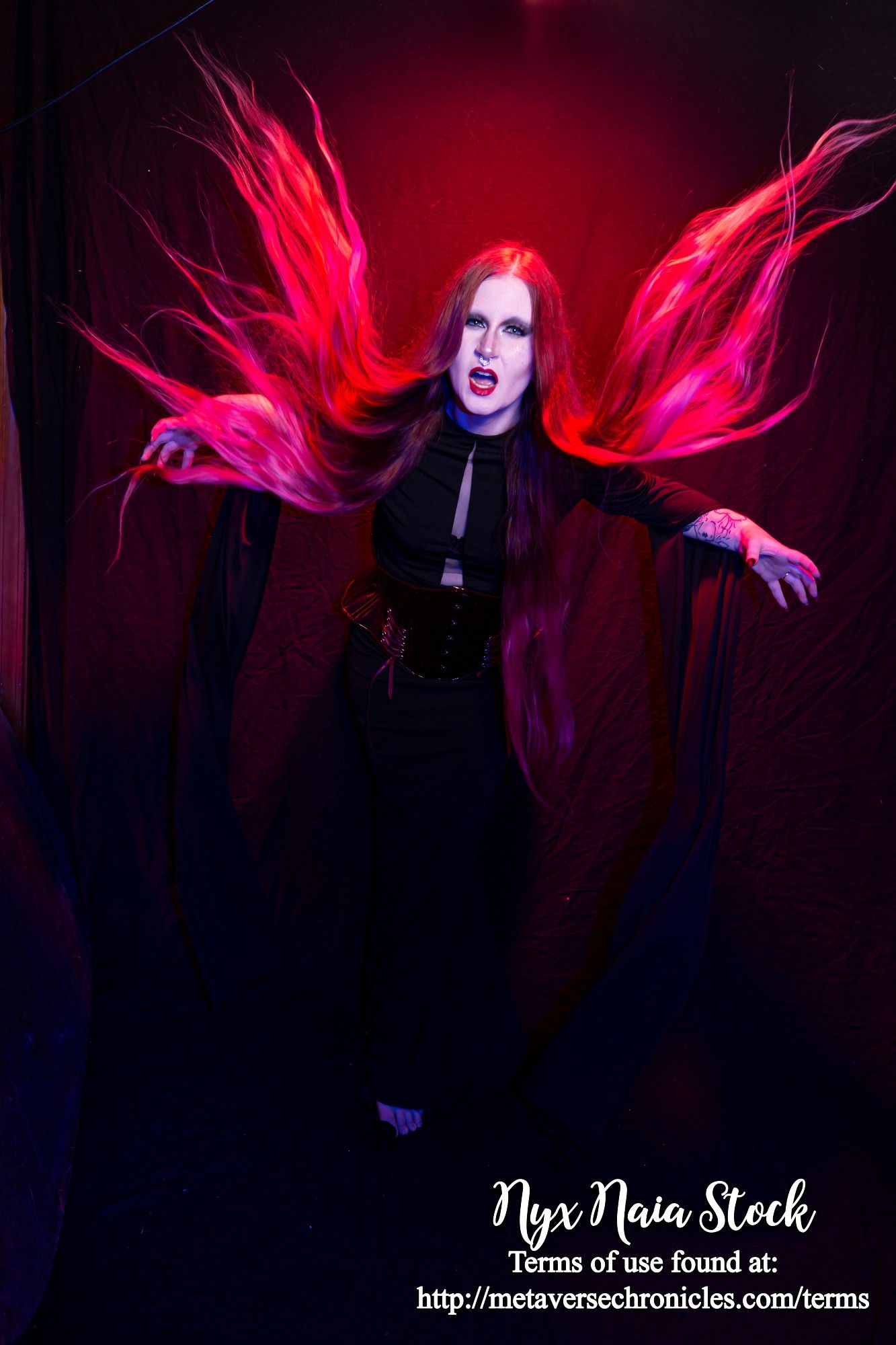 A photo of a white person with very long hair (Nyx Naia Stock) wearing a black long-sleeved dress. She has her arms out and is screaming at the camera, and her hair is flying around her, caught midmotion. Her hair is backlit by a red light, making it look like dancing flame.