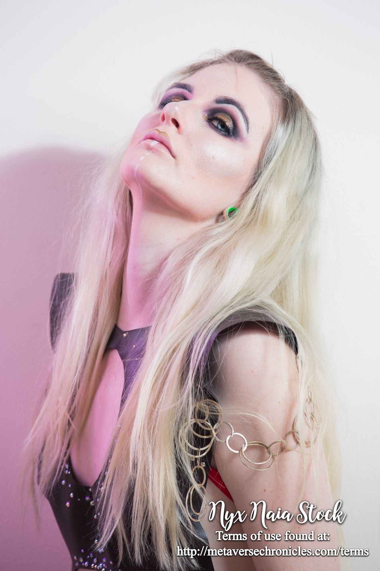 A photo portrait of a white person (Nyx Naia Stock) with medium-length blonde hair, wearing gold-and-black makeup and a sleeveless cutout shirt with rhinestones and chains hanging across the upper arms. Nyx Naia has her head tilted up and is looking down at the viewer.