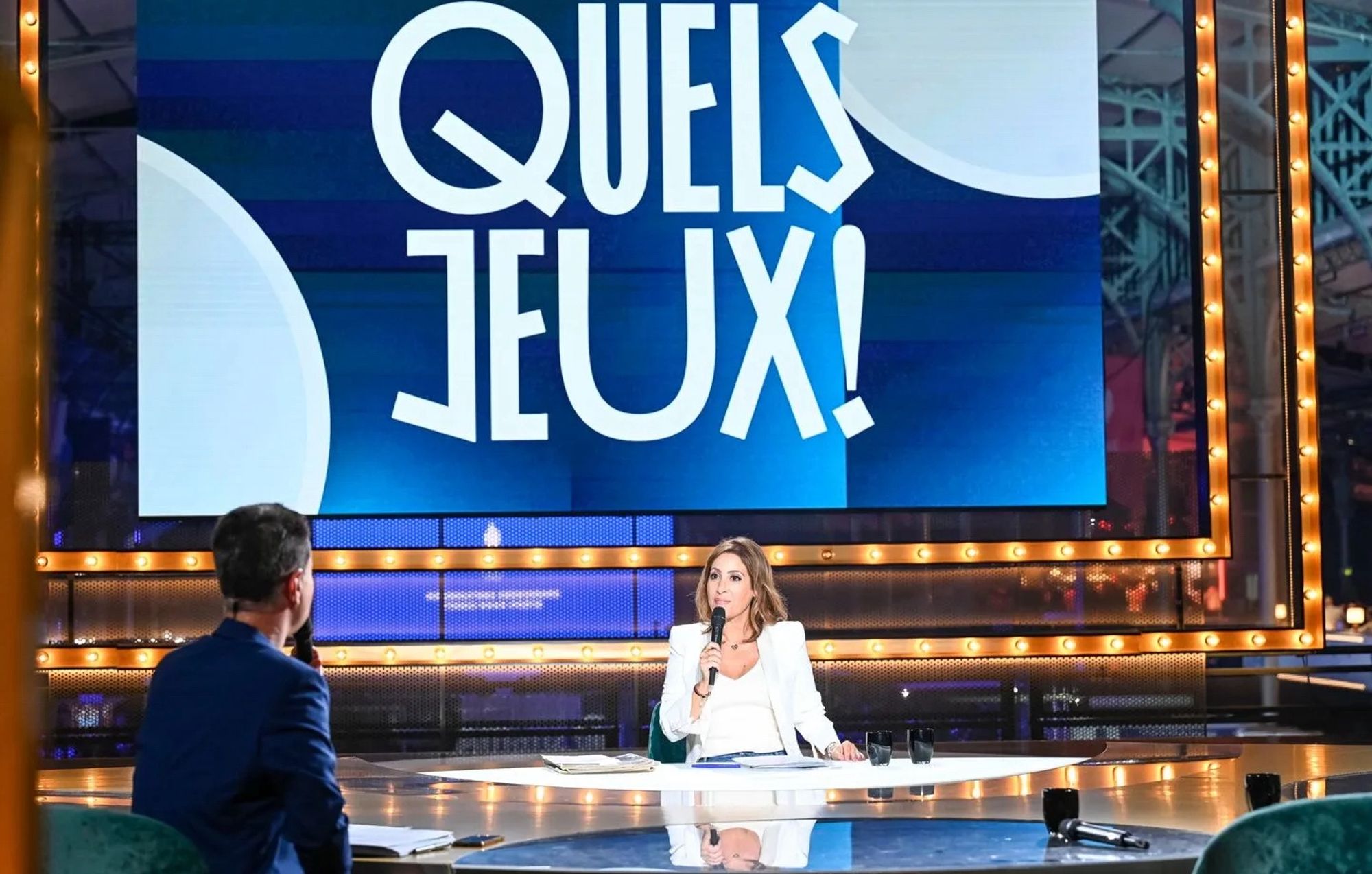 Léa Salamé vêtue de blanc est installée à la table sur le plateau de l’émission « Quels Jeux ! » dont le titre s’affiche en grand sur l’écran géant derrière elle. Elle s’adresse à Laurent Luyat qui est assis en face d’elle et que l’on voit ici de dos.