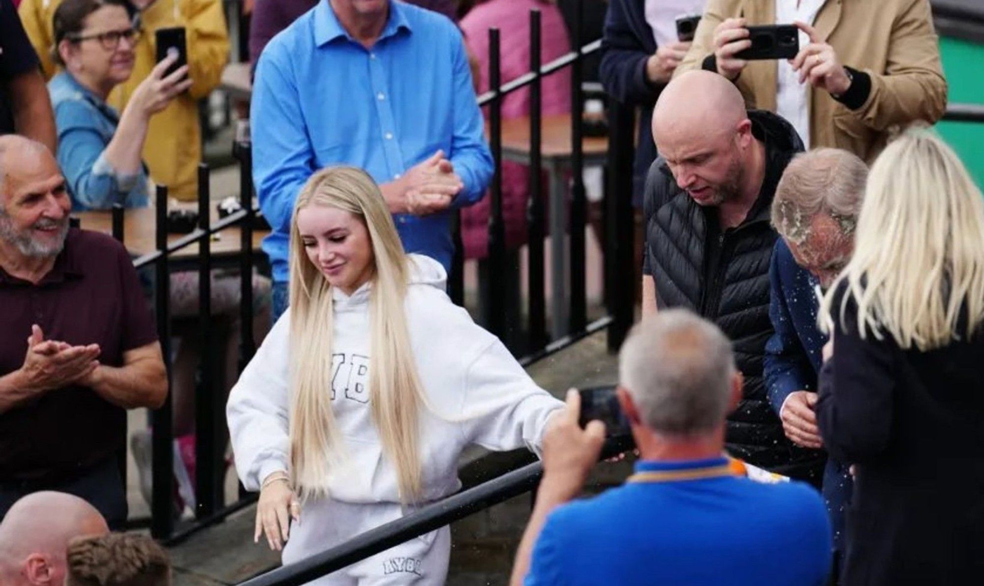 Person who threw milkshake on Nigel Farage walks away, looking pleased with herself, while audience smiles.