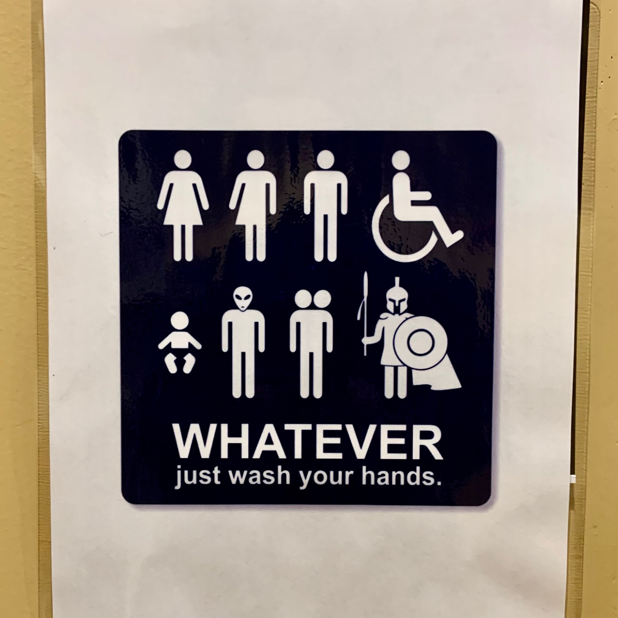 A sign depicting eight different figures, including stereotypical masc and fem figures, and a “hybrid” figure, and a figure in a wheelchair, and a figure with an alien head, and a figure with two heads, and a Valkyrie, and an infant. Phrase: “WHATEVER—just wash your hands.”