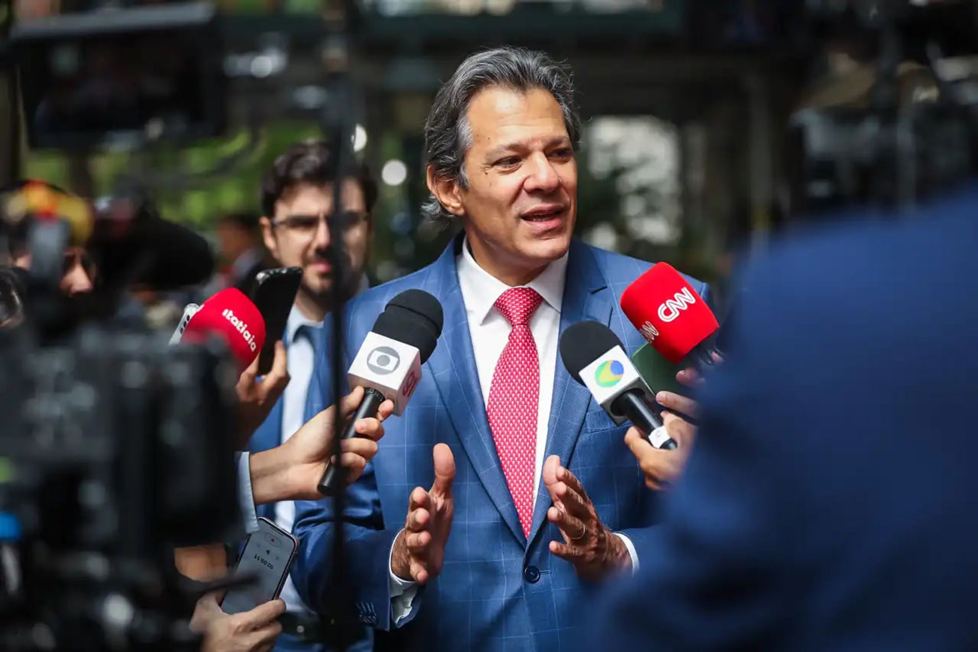 O ministro da Fazenda, Fernando Haddad, aparece em destaque na imagem, vestido com um terno azul e gravata vermelha de bolinhas brancas, cercado por microfones de diversas emissoras de TV, como Globo, CNN e BandNews. Ele gesticula enquanto fala com os jornalistas, sugerindo uma entrevista coletiva. Ao fundo, há pessoas desfocadas, uma delas segurando um celular, e uma leve paisagem externa.