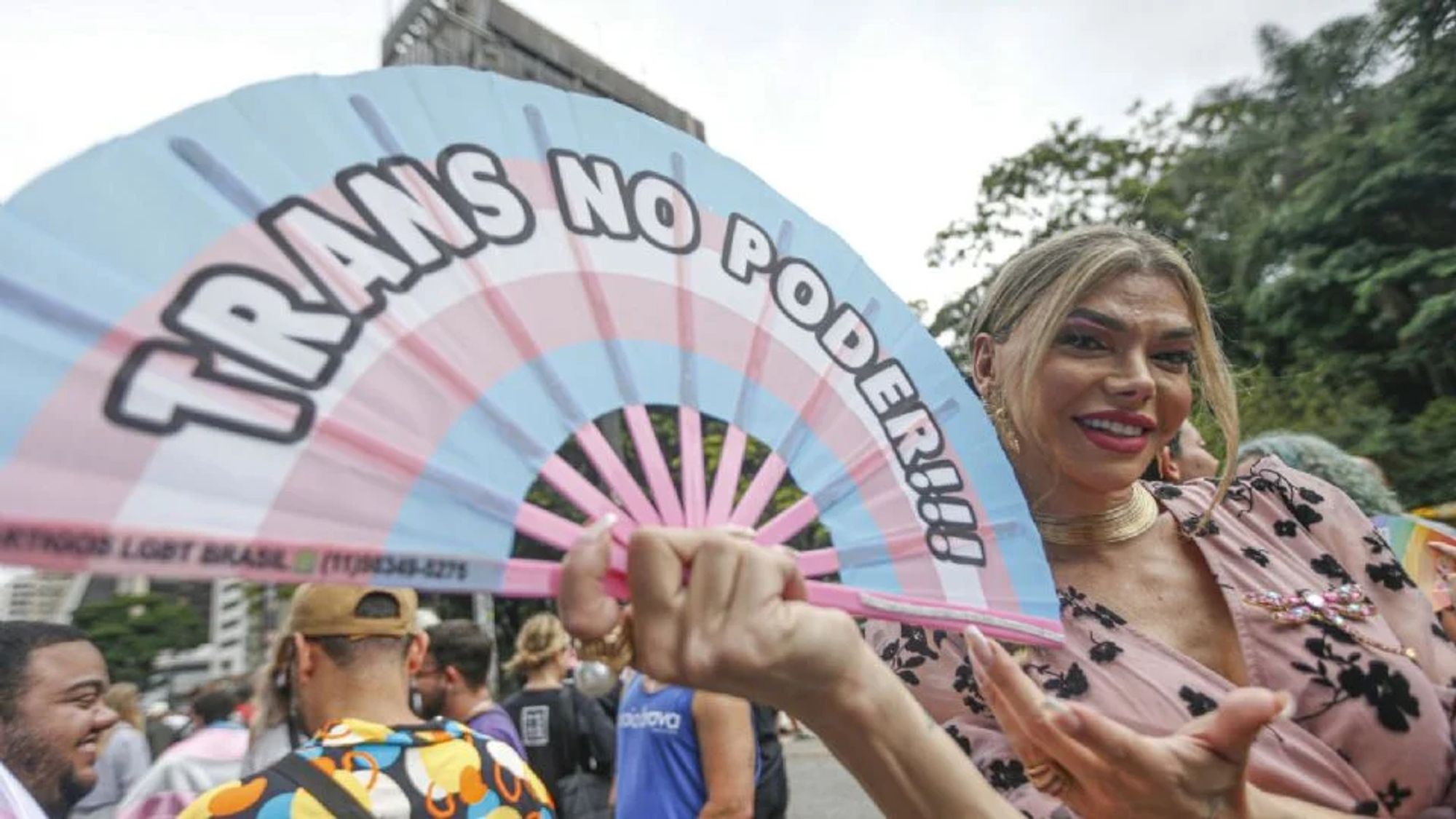 A imagem mostra uma pessoa participando da Caminhada Trans, em São Paulo, 2024, segurando um grande leque nas cores da bandeira transgênero (azul claro, rosa e branco), com a frase "TRANS NO PODER!!!" escrita em destaque. A pessoa sorri para a câmera, vestindo uma blusa rosa com detalhes coloridos. Ao fundo, é possível ver outros participantes e prédios típicos da Avenida Paulista. O evento celebra a visibilidade e os direitos da comunidade trans.