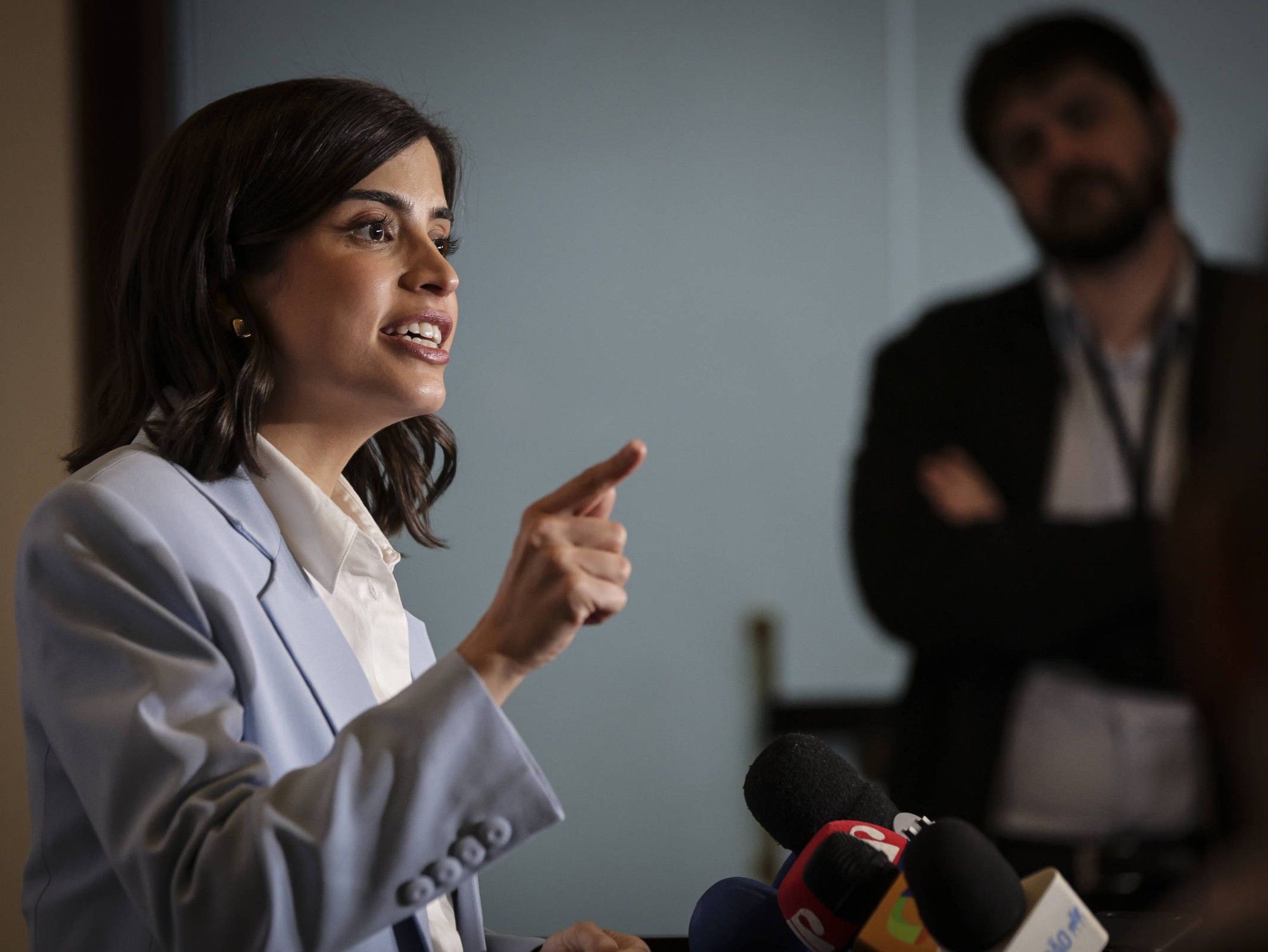 A imagem mostra a deputada federal Tabata Amaral falando durante uma coletiva de imprensa. Ela gesticula com a mão direita levantada e aponta com o dedo, expressando-se de forma enfática. Tabata veste um blazer azul-claro sobre uma camisa branca e está posicionada diante de vários microfones de diferentes veículos de comunicação. Ao fundo, desfocado, um homem de terno e barba observa com os braços cruzados.