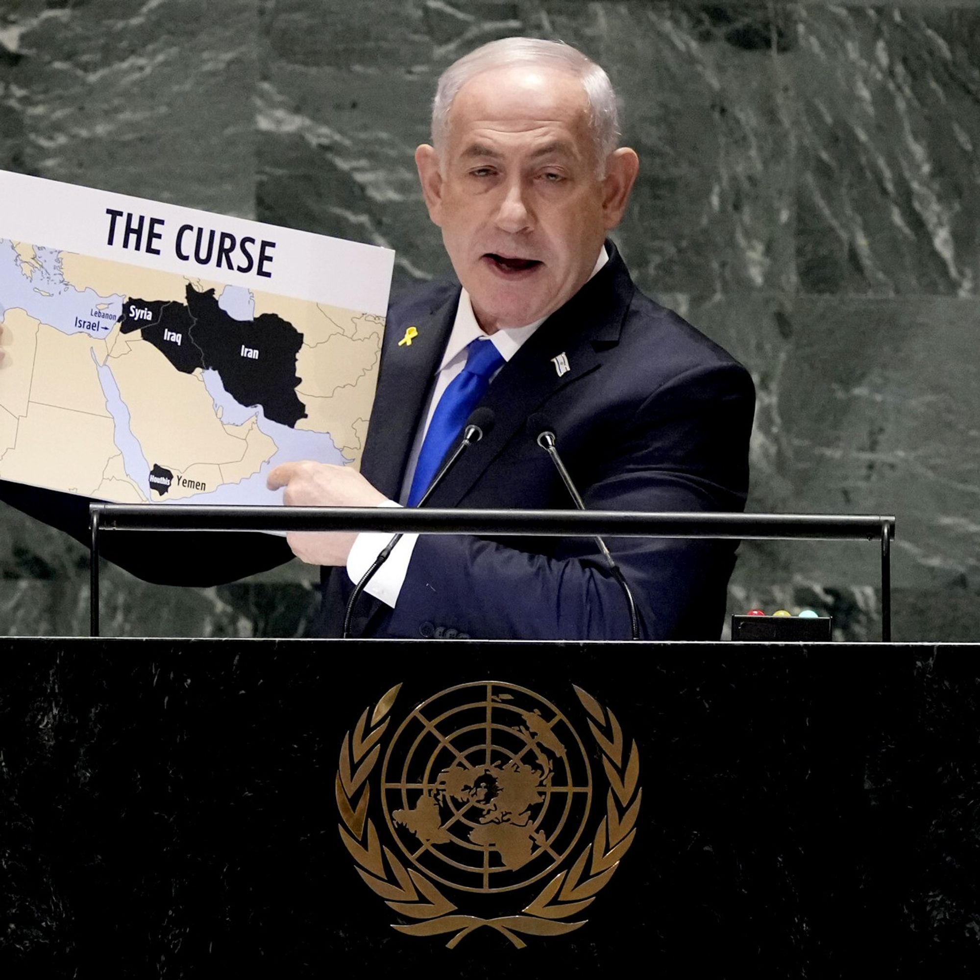 Benjamin Netanyahu, primeiro-ministro de Israel, é fotografado durante um discurso na Assembleia Geral da ONU. Ele segura um cartaz com o título "The Curse" ("A Maldição"), que mostra um mapa do Oriente Médio, destacando em preto países como Irã, Iraque, Síria e Iêmen. Netanyahu está de terno escuro com gravata azul e um pequeno laço amarelo preso à lapela. Diante dele, vê-se o emblema oficial da ONU.