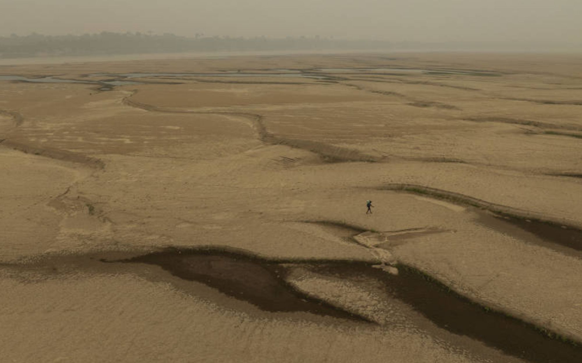 Uma vastidão seca e desolada, com o leito do rio Madeira praticamente sem água, revelando grandes áreas de solo exposto. Ao longe, um indivíduo caminha sozinho, dando a dimensão da imensidão do cenário árido. A paisagem está coberta por uma névoa acinzentada, sugerindo fumaça ou poluição no ar. Pequenos rastros de água aparecem em formações sinuosas no solo, mas são escassos e dispersos.