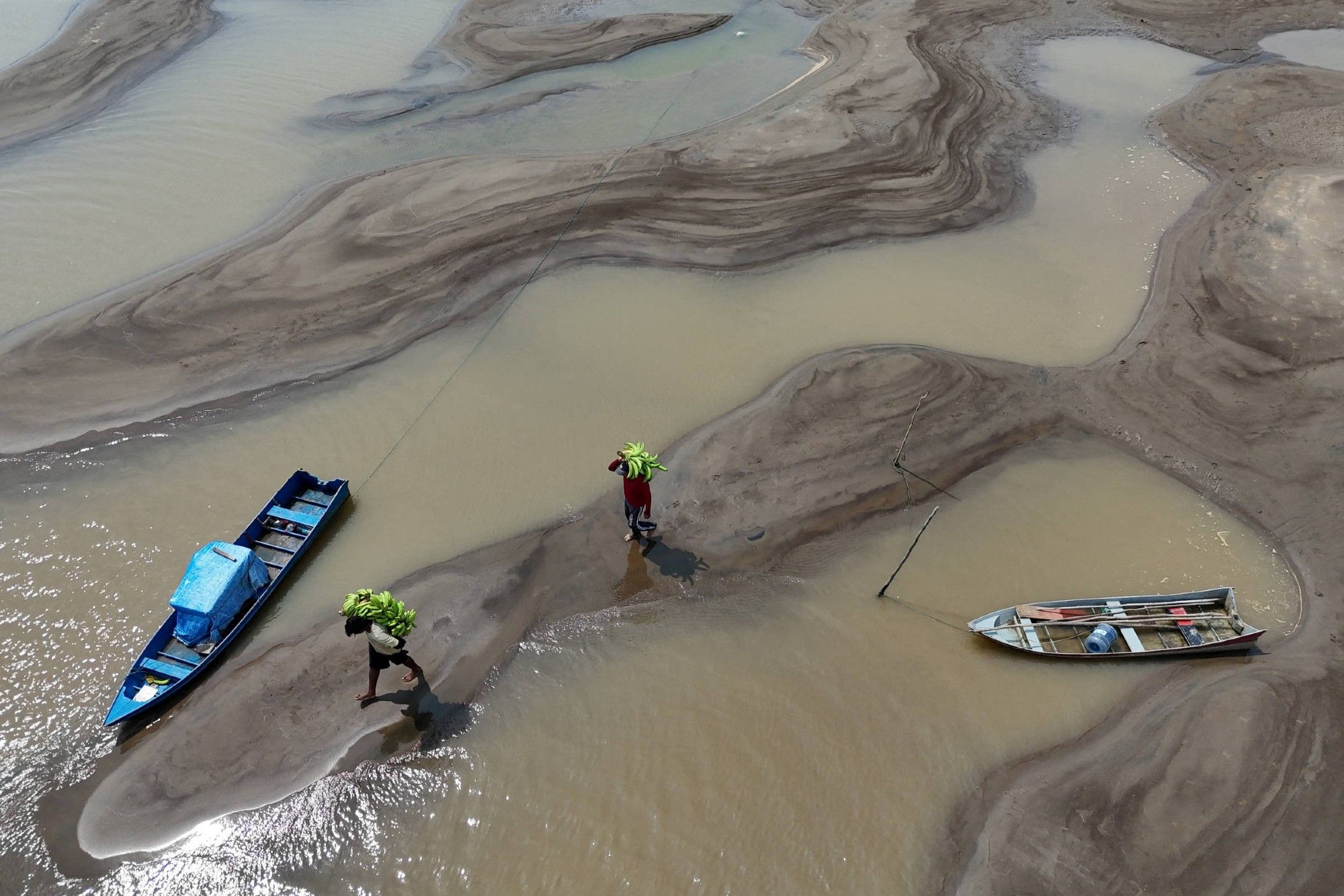 Imagem aérea do rio Solimões durante a seca, com vários bancos de areia à mostra. Duas pessoas carregam cachos de bananas, caminhando sobre os bancos de areia que agora dividem o rio em pequenos canais de água. Dois barcos, um azul e um de madeira, estão encalhados nas áreas de areia, evidenciando o nível crítico da água. A paisagem parece caótica, com curvas sinuosas de lama e água formando padrões irregulares.