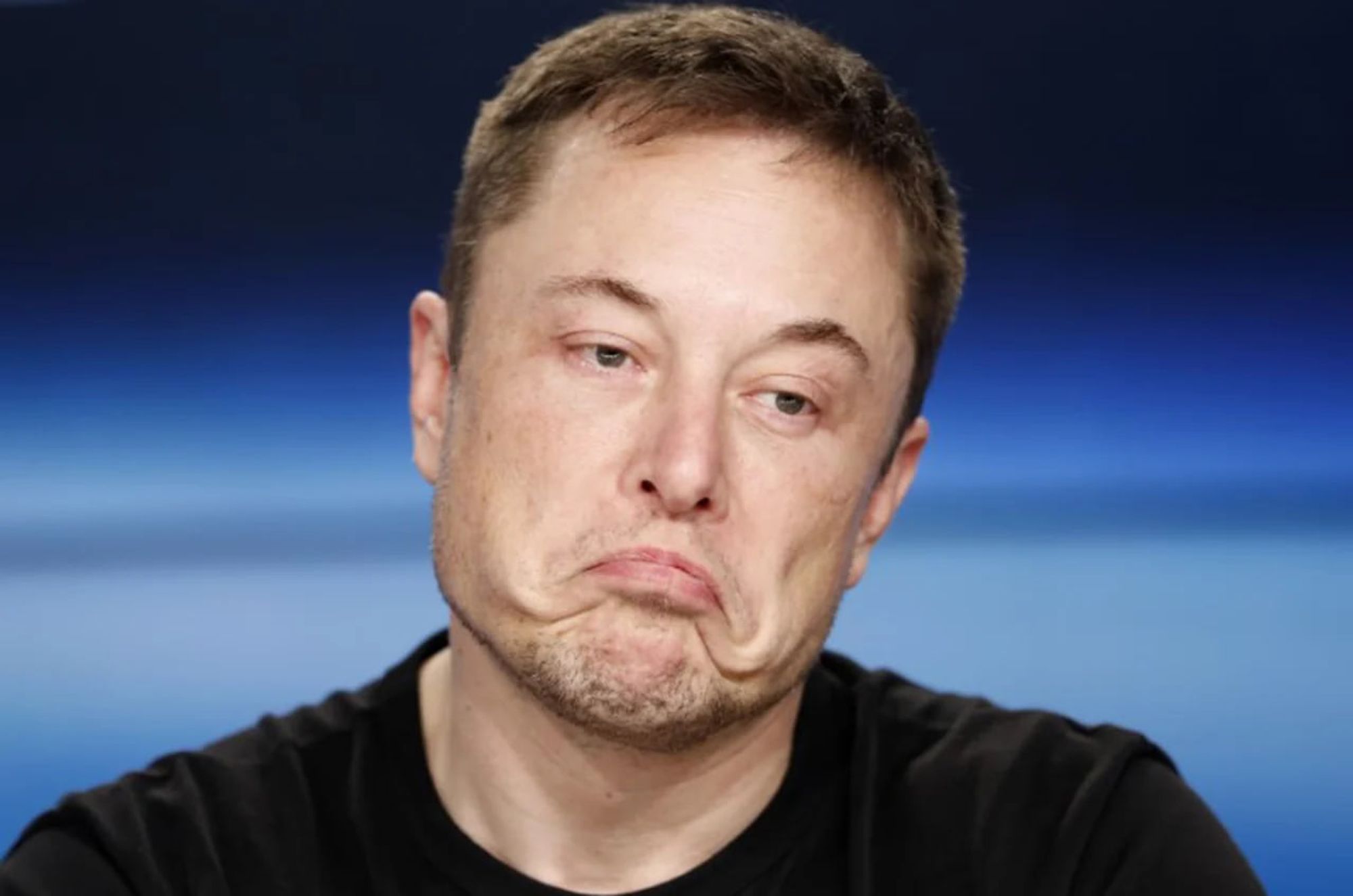 Imagem de Elon Musk fazendo uma expressão facial de desdém ou indiferença, com os lábios curvados para baixo de forma exagerada. Ele veste uma camiseta preta, e o fundo desfocado tem um tom de azul gradiente. A foto captura um momento informal ou descontraído do empresário.