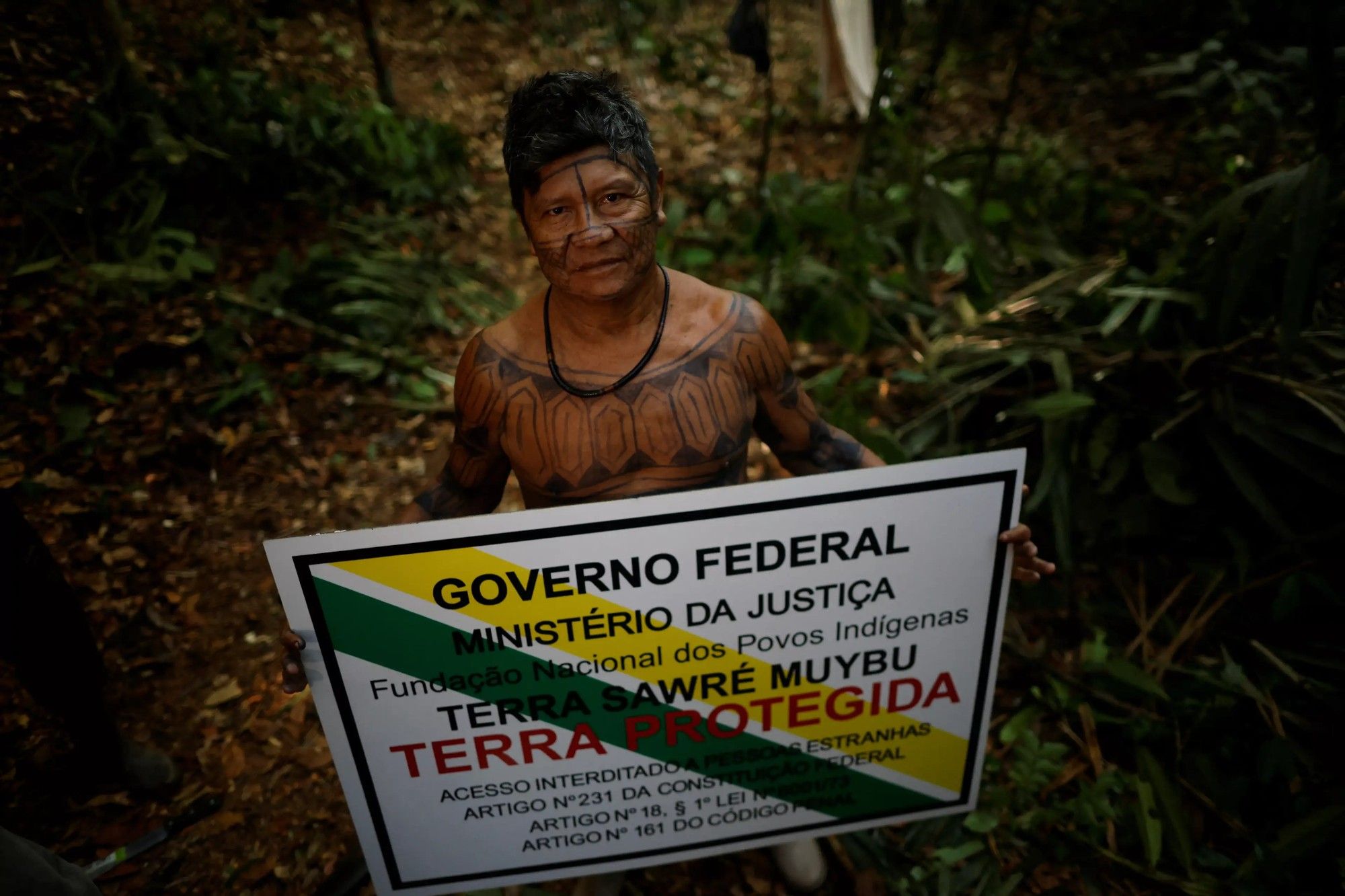 Imagem do cacique Juarez Saw, da aldeia Sawré Muybu, segurando uma placa que indica a demarcação de território indígena em Itaituba, no estado do Pará. Ele está em pé em uma área de floresta, com o corpo pintado com desenhos geométricos tradicionais. A placa que ele segura traz as seguintes inscrições: "Governo Federal, Ministério da Justiça, Fundação Nacional dos Povos Indígenas, Terra Sawré Muybu, Terra Protegida. Acesso interditado a pessoas estranhas. Artigo nº 231 da Constituição Federal, Artigo nº 18, § 1º Lei nº 9.605/98, Artigo nº 161 do Código Penal."