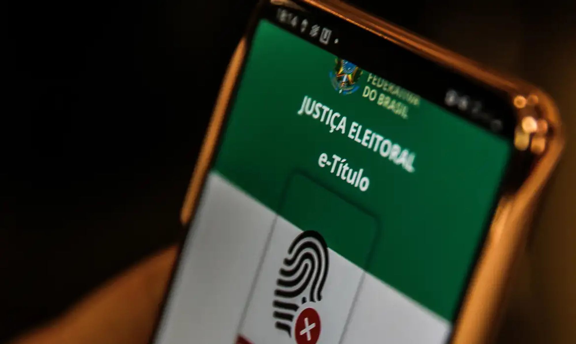 Foto de um smartphone mostrando a tela do aplicativo e-Título da Justiça Eleitoral do Brasil. Na parte superior da tela, há o brasão da República com a inscrição "Federação do Brasil" e abaixo o texto "Justiça Eleitoral" e "e-Título" em branco sobre um fundo verde. Abaixo disso, há a imagem de uma impressão digital com um "X" vermelho sobreposto, indicando possível erro ou necessidade de autenticação.