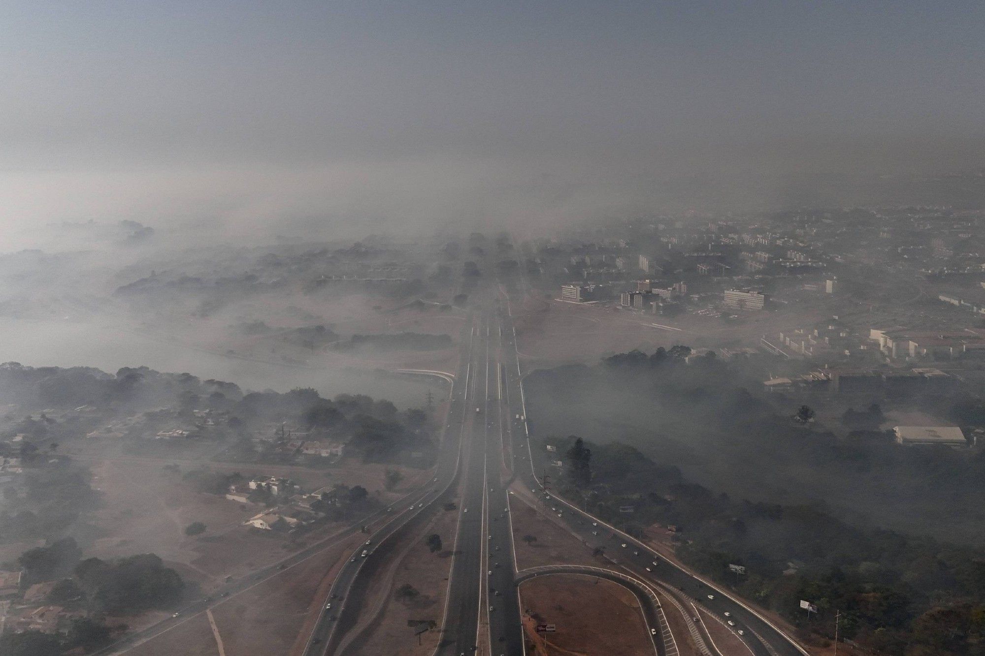 Vista aérea da Asa Norte, em Brasília, encoberta por uma densa camada de fumaça. A imagem mostra uma longa avenida, com carros trafegando, que se estende em direção ao horizonte, onde a fumaça se mistura ao céu, obscurecendo os edifícios e vegetação. A fumaça reduz a visibilidade, criando um cenário sombrio, com a cidade parcialmente coberta. A poluição é evidente, afetando tanto as áreas urbanas quanto os arredores mais arborizados.