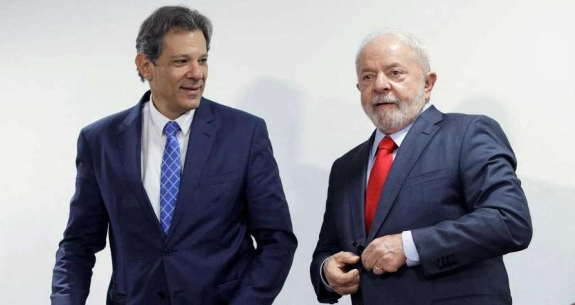 A imagem mostra Fernando Haddad e Luiz Inácio Lula da Silva lado a lado, ambos vestidos com ternos escuros. Haddad, à esquerda, usa uma gravata azul com padrões geométricos e sorri levemente enquanto olha para Lula. Lula, à direita, está usando uma gravata vermelha e parece ajustar o botão de seu paletó enquanto olha ligeiramente para a câmera. Ambos estão em um ambiente interno com um fundo neutro e claro.