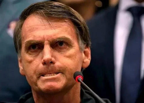Close de Jair Bolsonaro com expressão tensa, mordendo o lábio inferior e olhando fixamente para frente, como se estivesse segurando um comentário atravessado.