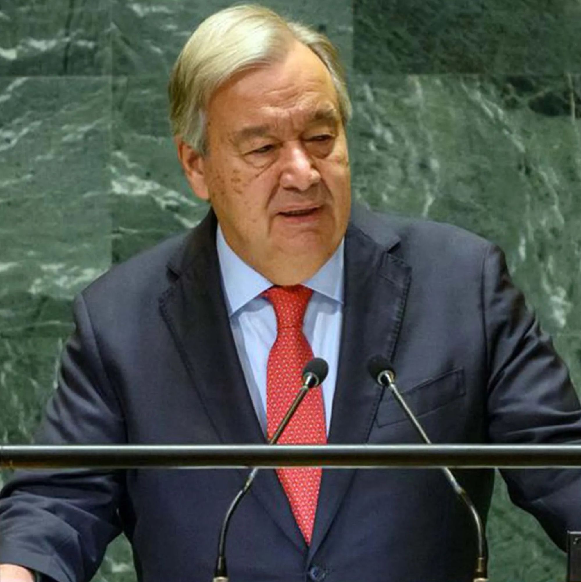 António Guterres, Secretário-Geral das Nações Unidas, discursa em um púlpito com dois microfones, vestido com terno escuro, camisa branca e gravata vermelha. Ele está em frente a um fundo verde com padrões de mármore, que é característico da sede da ONU. Guterres parece sério e focado durante o discurso.