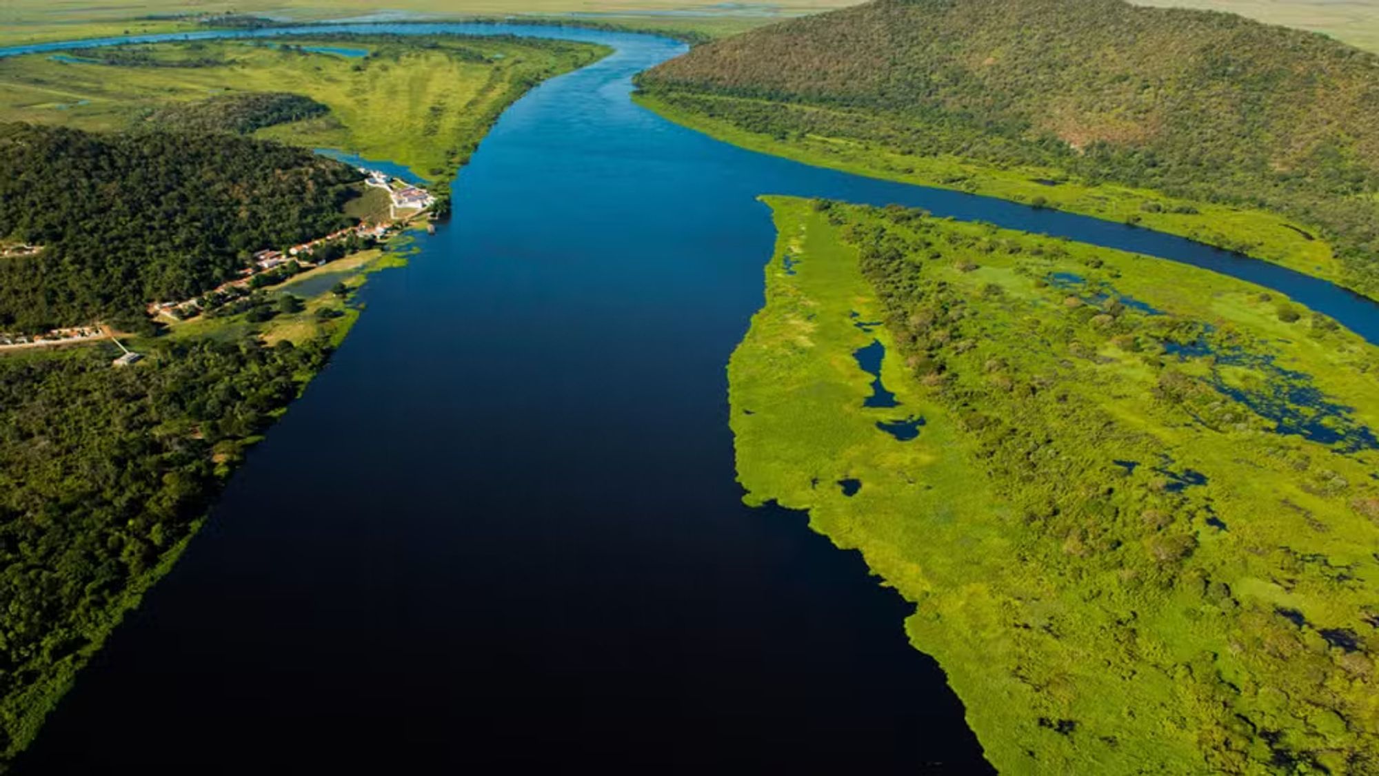 Imagem aérea da Bacia do Alto Paraguai, no Pantanal mato-grossense. O rio serpenteia pelo centro da imagem, com águas escuras que contrastam com a vegetação verde exuberante que o cerca. À esquerda, há uma faixa de floresta mais densa, com algumas construções visíveis ao longo da margem do rio. À direita, uma planície alagada com vegetação rala e diversos pequenos corpos d'água. Ao fundo, avistam-se campos e morros que se estendem até o horizonte, caracterizando a vasta paisagem natural do Pantanal.