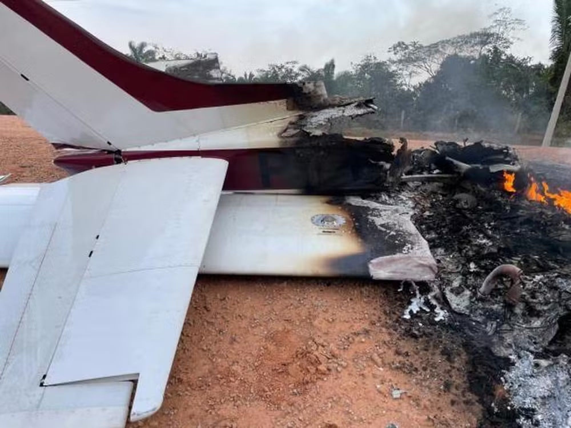 Imagem de uma aeronave parcialmente destruída pelo fogo. A parte traseira da fuselagem e a cauda da aeronave, com listras em vermelho e branco, ainda estão inteiras, mas a parte central está queimada e derretida. À direita, chamas ainda estão visíveis, consumindo o que restou do avião. A aeronave está sobre um terreno de terra batida e há árvores ao fundo, sugerindo que o incidente ocorreu em uma área remota.