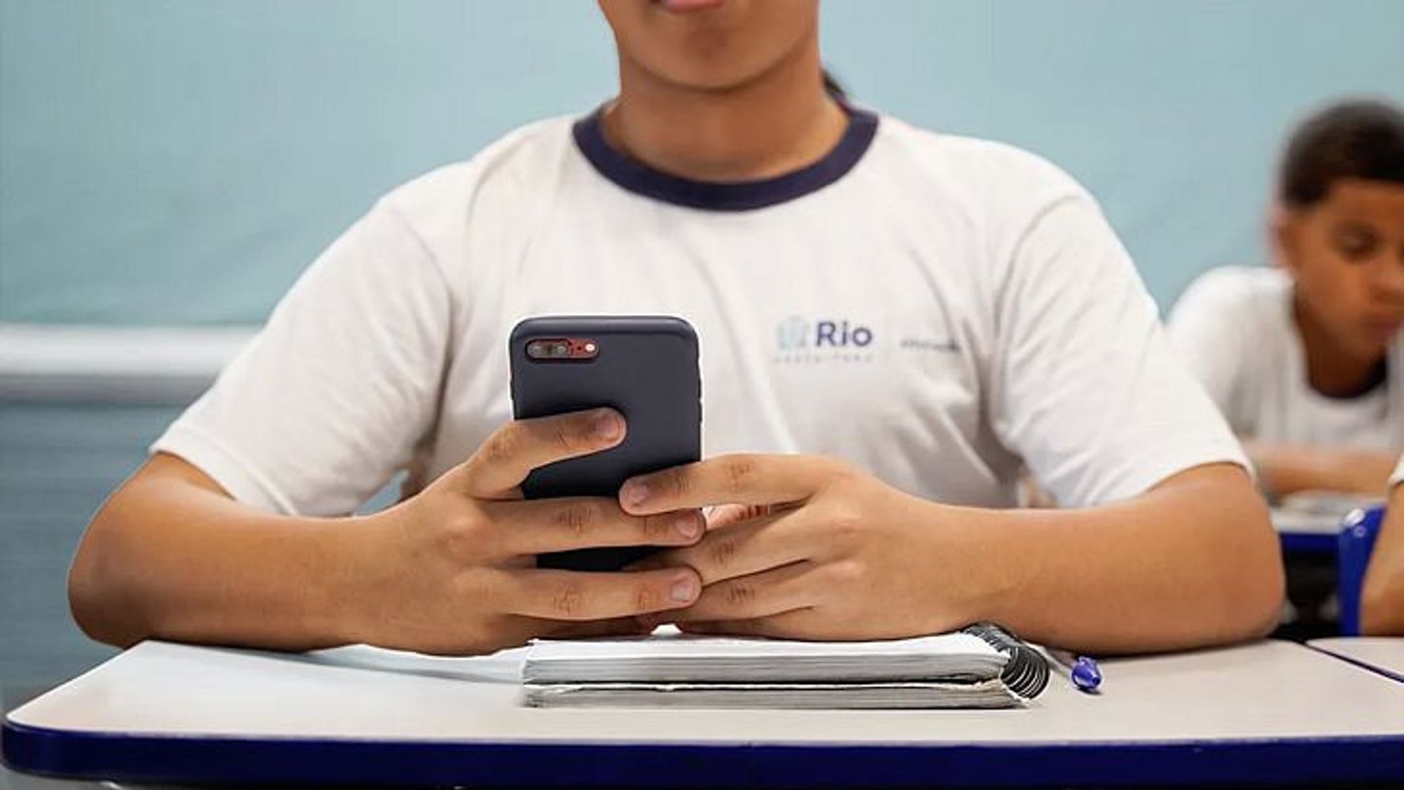 Imagem de um estudante sentado em uma carteira escolar, vestindo uniforme branco com detalhes em azul e o logo da Prefeitura do Rio de Janeiro estampado na camisa. Ele segura um celular com as duas mãos, enquanto um caderno e uma caneta estão sobre a mesa à sua frente. Ao fundo, outro aluno aparece desfocado, sugerindo um ambiente de sala de aula.