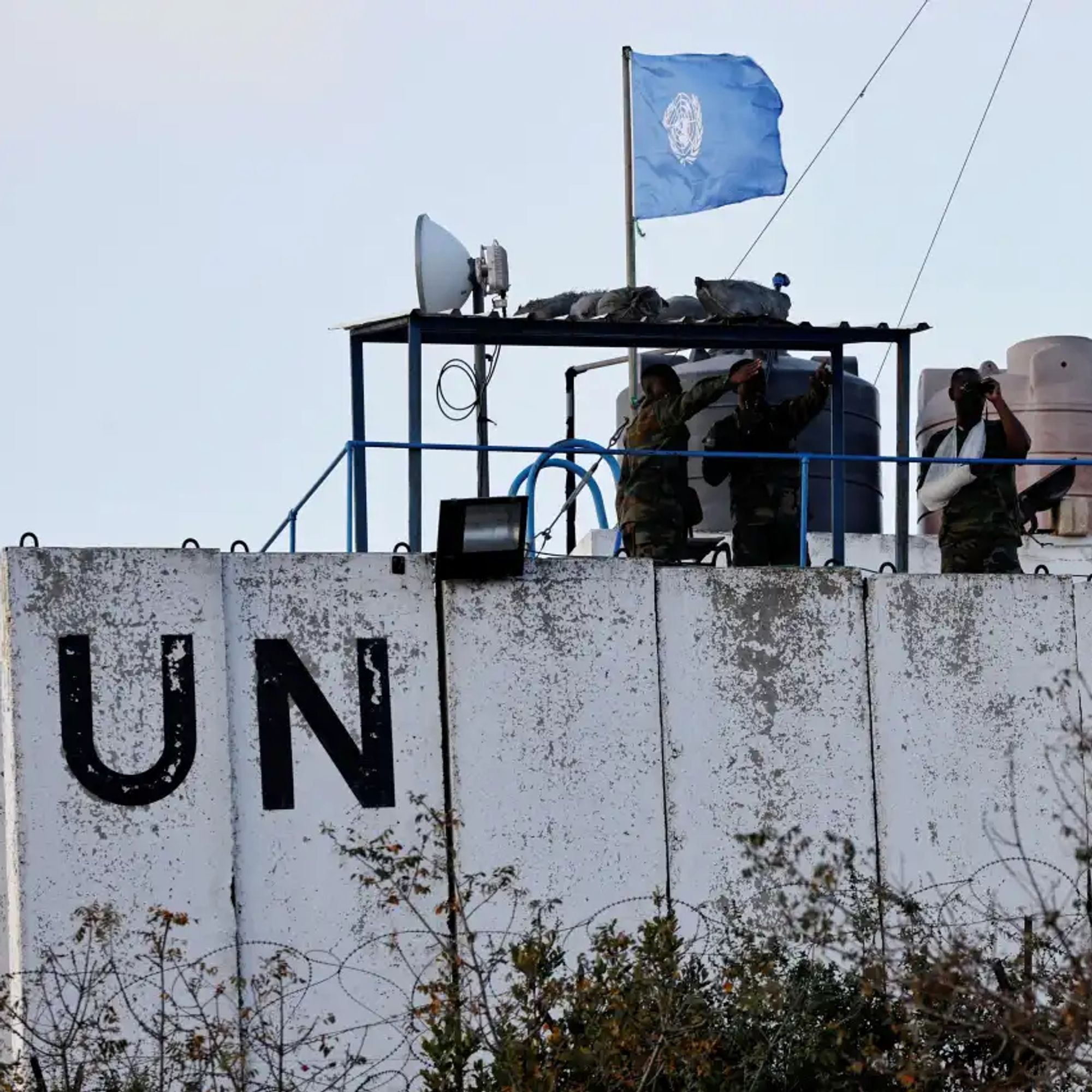 A imagem mostra três soldados da missão de paz da ONU em uma estrutura elevada na fronteira entre Líbano e Israel. Eles estão uniformizados e observam a área ao redor, com um deles utilizando binóculos. A estrutura tem uma bandeira azul das Nações Unidas hasteada e a parede da construção exibe as letras "UN" em preto. O local é cercado por antenas e equipamentos de comunicação. A paisagem ao redor apresenta arbustos e cercas de arame, reforçando a sensação de uma zona de fronteira vigiada.