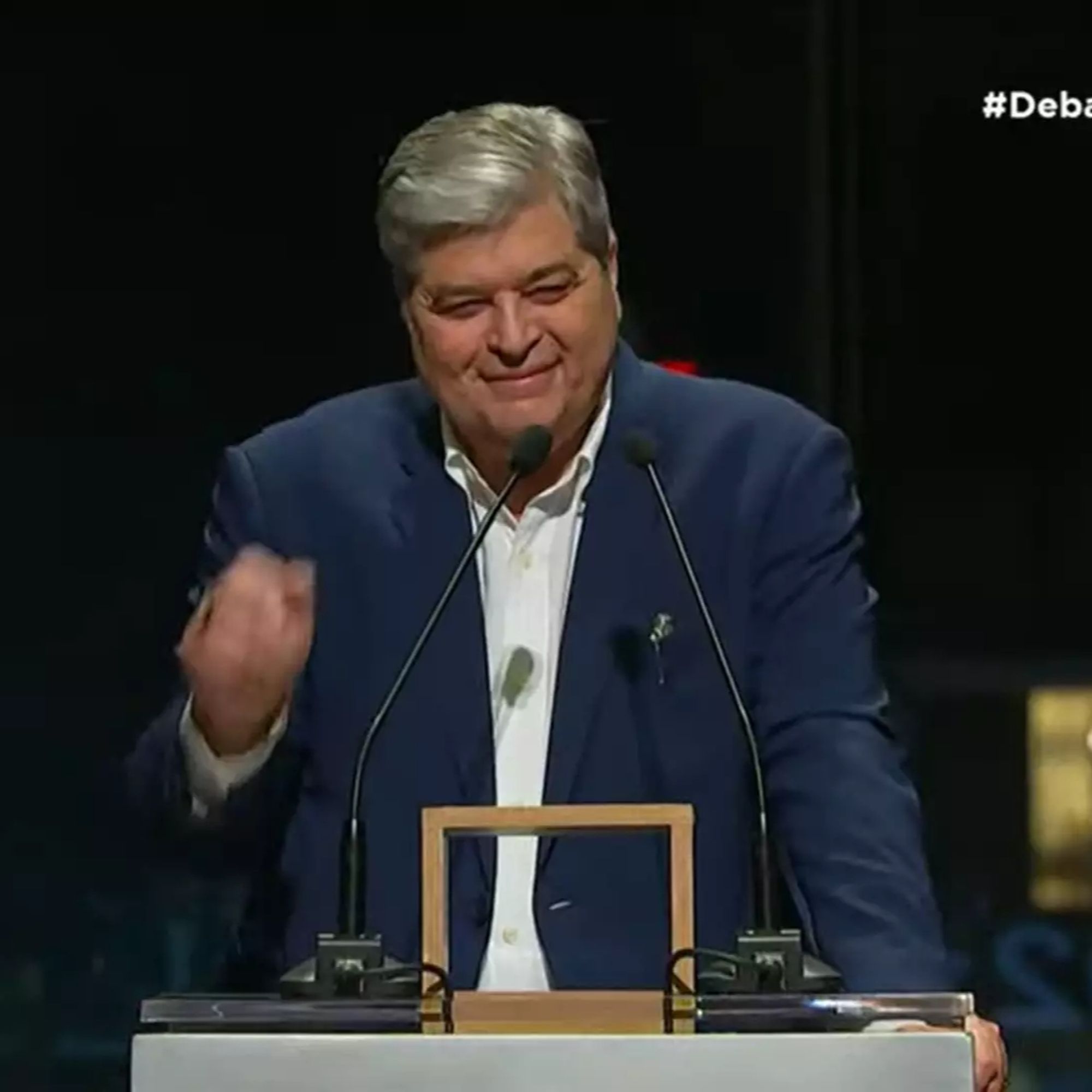 Imagem de José Luiz Datena durante o debate da TV Cultura. Ele está em pé atrás de um púlpito, com dois microfones à sua frente. Datena usa um terno azul escuro e camisa branca, e faz um gesto com a mão direita. Ele sorri de forma discreta enquanto olha para a frente. O fundo é escuro, destacando o apresentador, e no canto superior direito da imagem aparece a hashtag "#Debate".