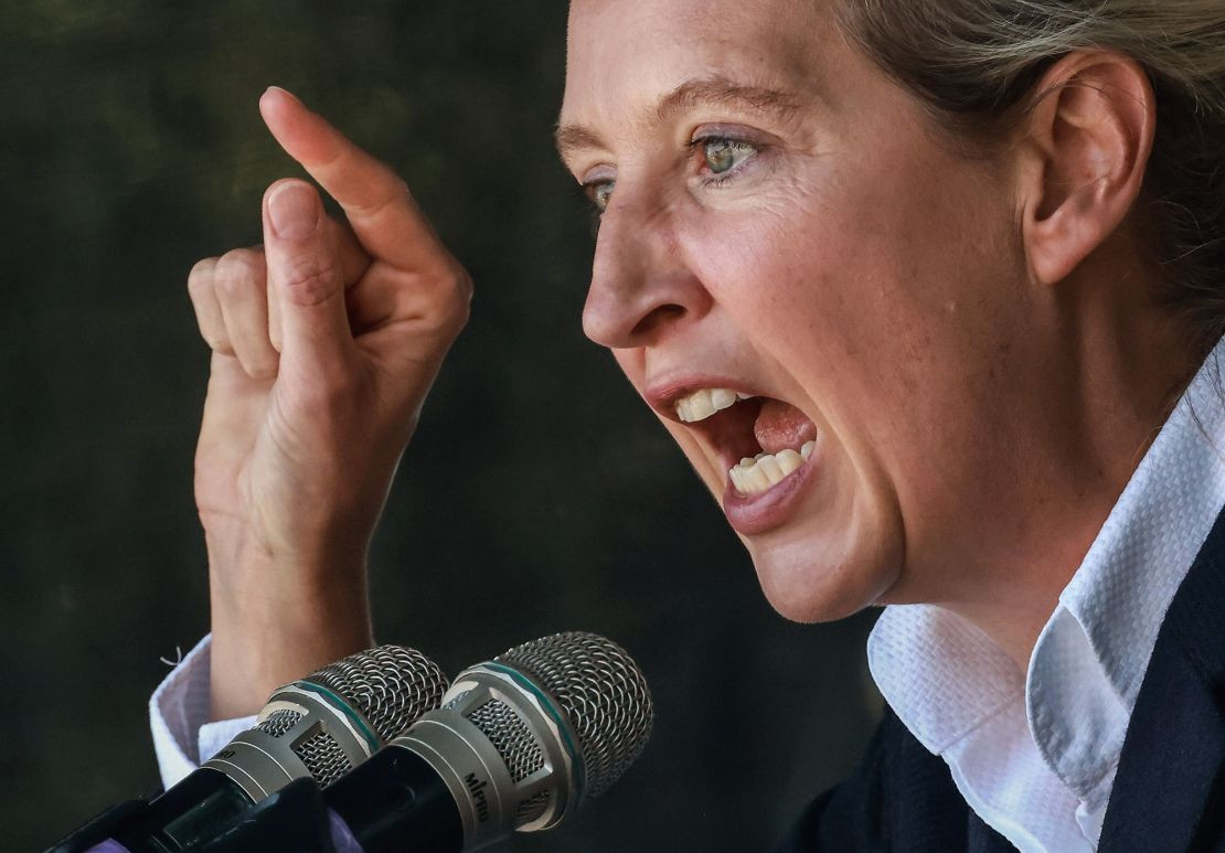 A imagem mostra Alice Weidel, co-líder do partido AfD, em um momento de discurso enérgico. Ela está falando com intensidade ao microfone, com expressão séria e gesticulando com a mão direita, apontando para cima. Weidel veste uma camisa branca e está de perfil, evidenciando sua postura firme e assertiva.