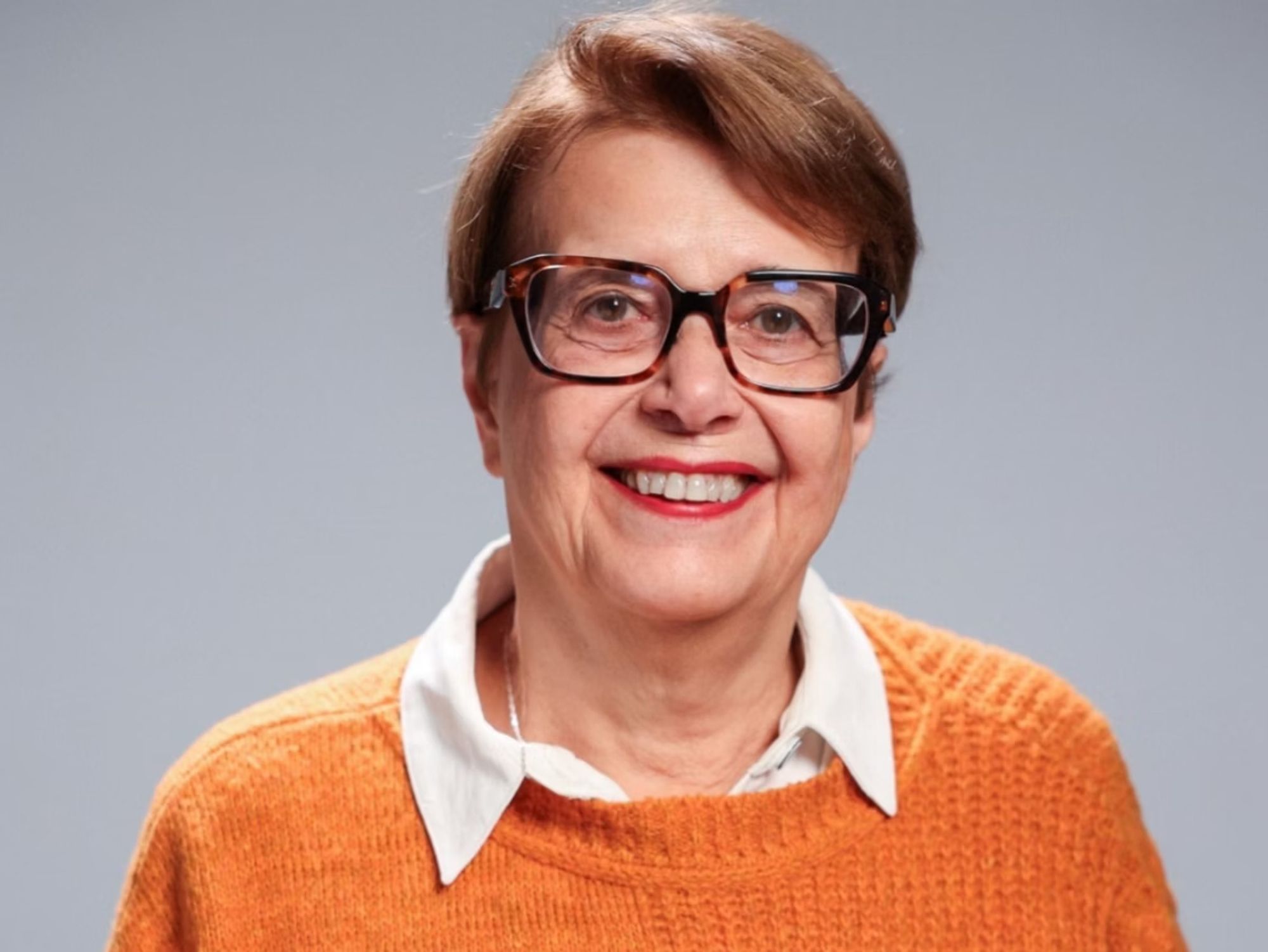 A imagem mostra uma mulher de pele clara, cabelos curtos castanhos e usando óculos de armação preta e grossa. Ela está sorrindo e veste uma blusa laranja por cima de uma camisa branca. O fundo é simples, com uma cor sólida em tom cinza. A legenda indica que se trata de Margarida Salomão, prefeita de Juiz de Fora, que busca reeleição. A foto foi compartilhada em seu Instagram.