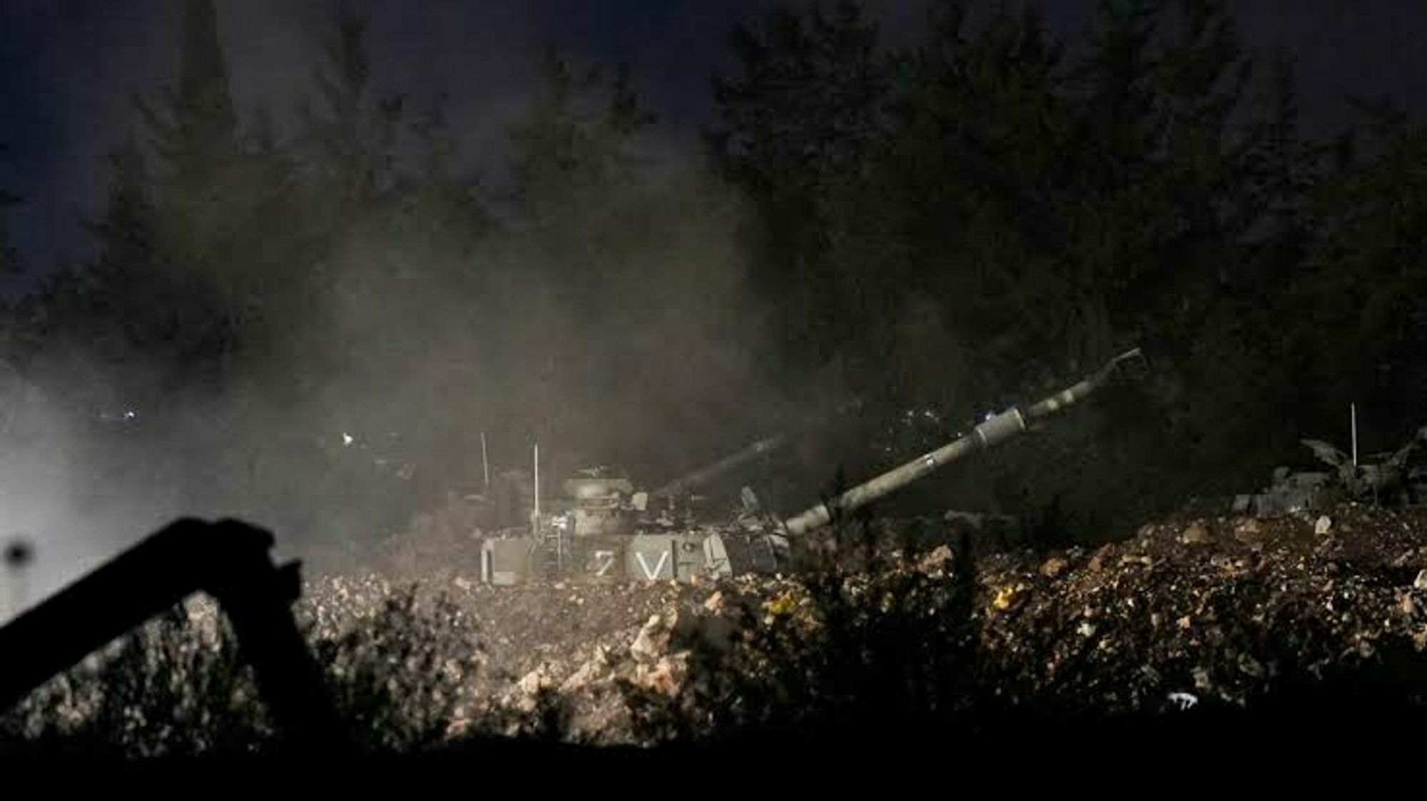 An Israeli mobile artillery unit is positioned on rocky terrain in northern Israel, firing a shell towards Lebanon. The scene is captured at night, with smoke and dust rising from the area. The vehicle is partially obscured by darkness and surrounding trees, and a "V" marking is visible on its side. The photograph depicts military action near the Israel-Lebanon border on September 30.