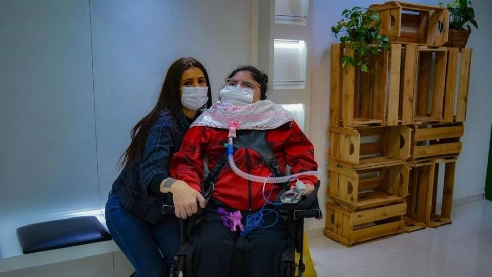 A imagem mostra duas mulheres em um ambiente interno. À esquerda, Luciana Nietto, de cabelo longo e escuro, vestindo uma jaqueta jeans, está abraçando a filha à direita, que está em uma cadeira de rodas. Heloísa Nietto usa uma máscara de oxigênio e uma roupa vermelha, parece ter necessidades especiais e está conectada a equipamentos médicos, incluindo um respirador. Ambas estão usando máscaras faciais. Ao fundo, há uma estante feita de caixotes de madeira com algumas plantas no topo, e o ambiente é bem iluminado, com paredes brancas e decoração minimalista.