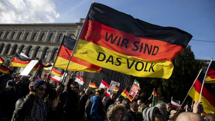 A imagem mostra uma manifestação do AfD na Alemanha com várias pessoas carregando bandeiras do país. Uma das bandeiras principais tem a frase "Wir sind das Volk" (Nós somos o povo) escrita sobre as cores da bandeira alemã. A multidão parece estar em uma praça pública, com muitos manifestantes segurando cartazes e bandeiras.