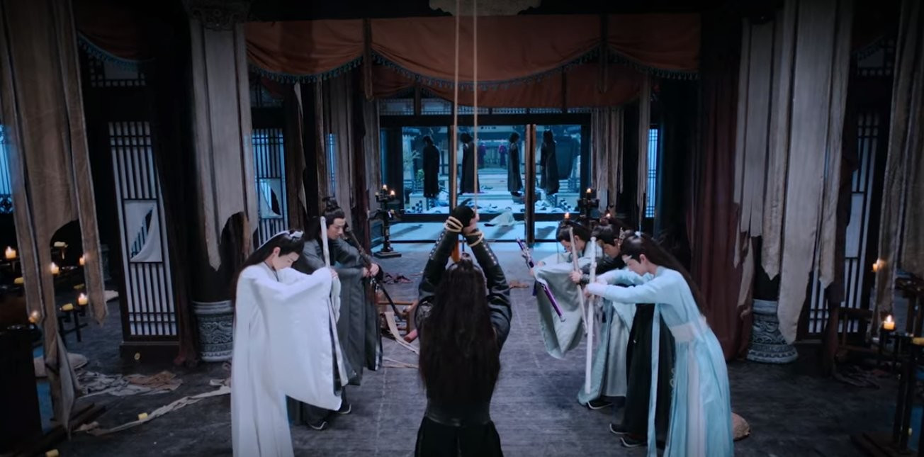 Wide shot of a room in Chang Manor. At the head of the room, opposite the door and closest the viewer, is Xue Yang, shown from behind, hands bound together above his head with cords hanging from the offscreen ceiling. Beyond Xue Yang a group of cultivators are facing each other in two rows on either side of Xue Yang, Xiao Xingchen and Song Lan to the left and Lan Wangji, Wei Wuxian, and Jiang Cheng on the right, bowing to those in the opposite row.