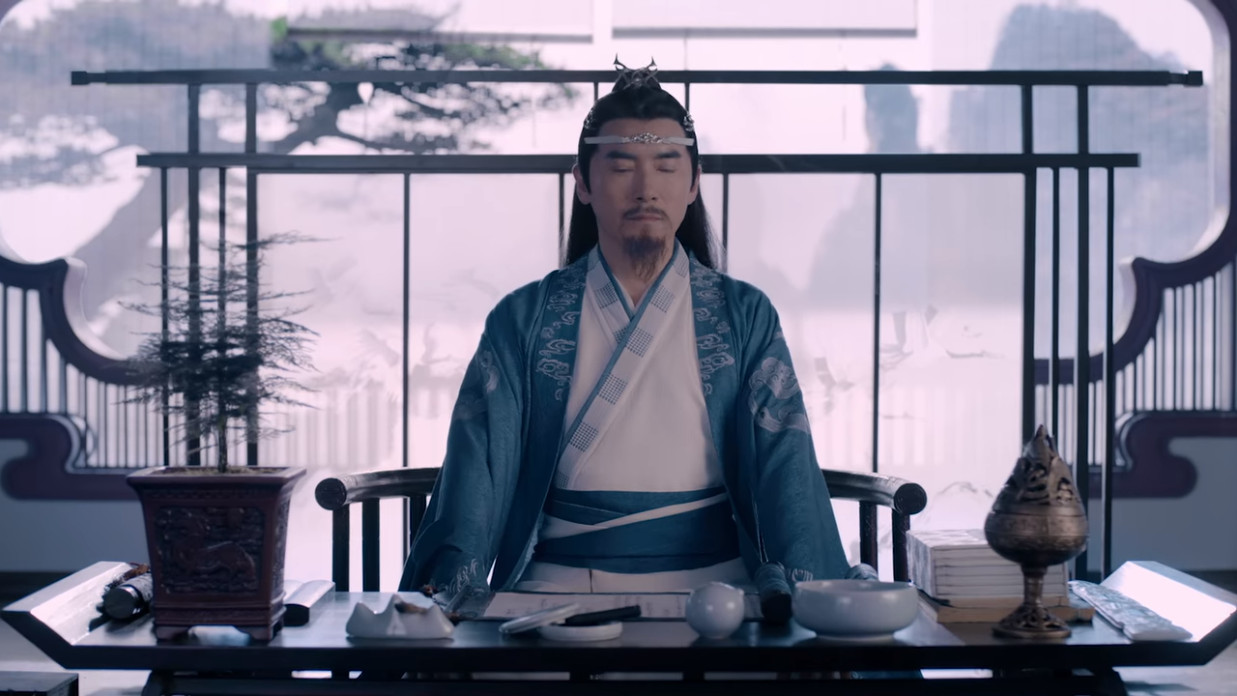 Lan Qiren sits behind a desk, and in front of a window, eyes closed in what could either be meditation or an attempt at escaping at whatever else might be going on in the room offscreen.