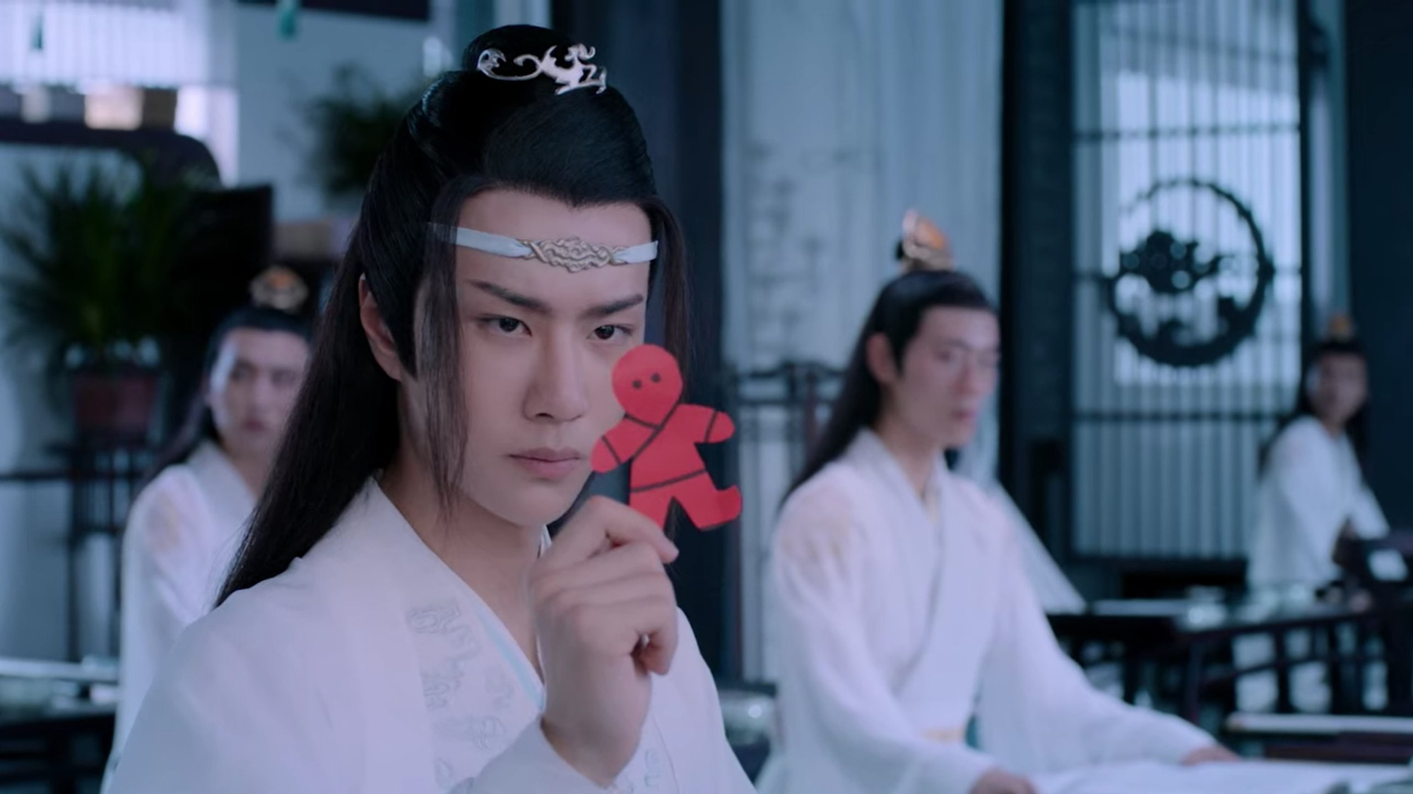 In a classroom during the lectures at the Cloud Recesses, teen Lan Wangji holds up a red paperman marked with black lines to indicate clothing boundaries in front of his face and studies it.