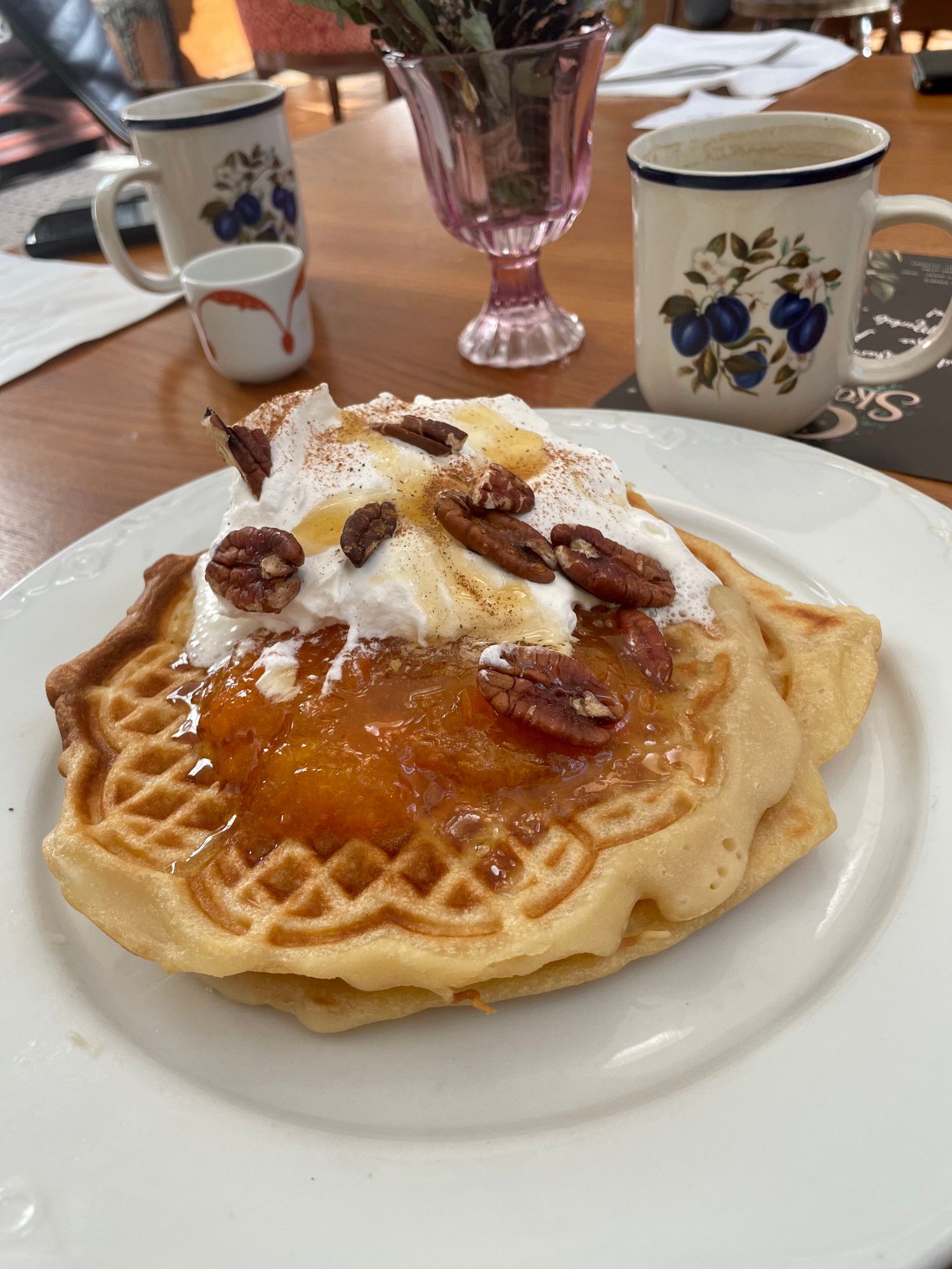 Two vegan, gluten-free waffles on a white plate. The waffles are topped with pumpkin jam, vegan whipped cream, pecans, cinnamon, and pumpkin drizzle. In the background are coffee cups.